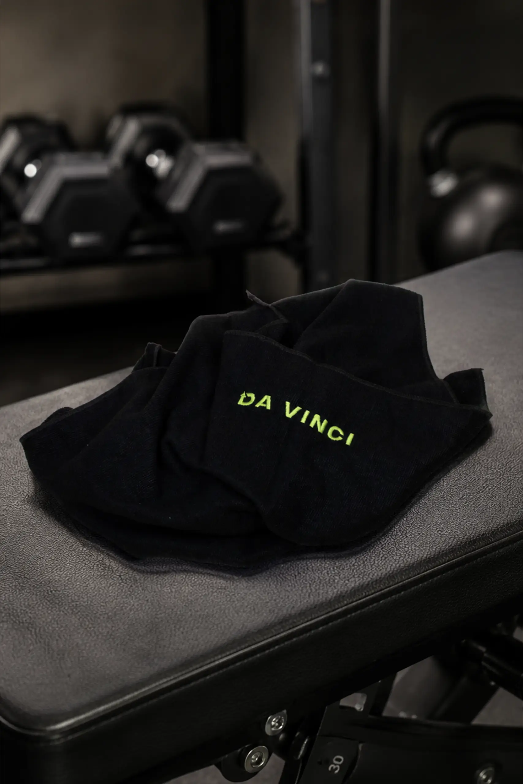 Folded black towel with the "Da Vinci" logo embroidered in lime green, placed on a gym bench with weights and dumbbells in the background.