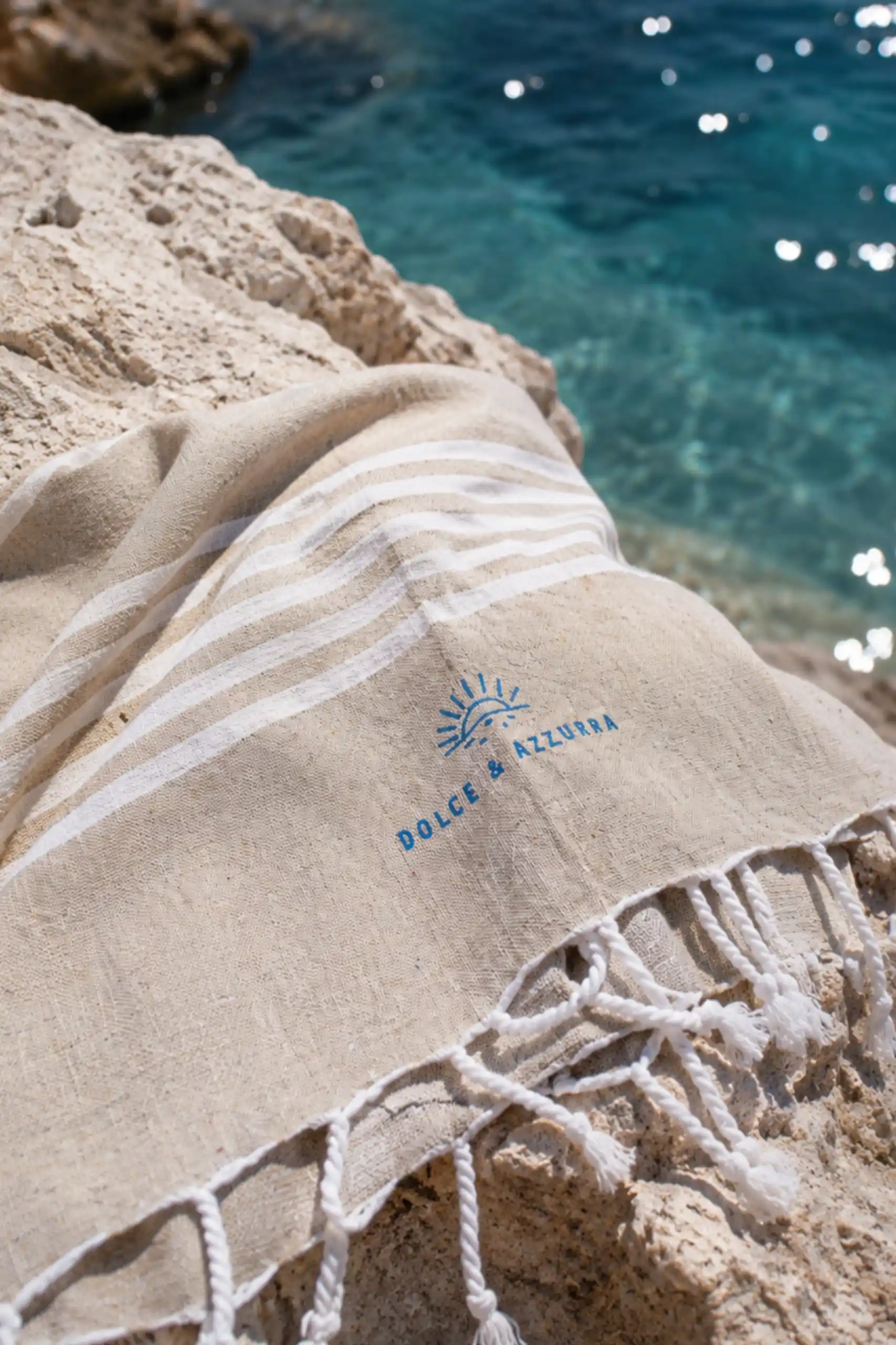 Primer plano de una toalla de playa color arena con rayas blancas y el logo bordado 