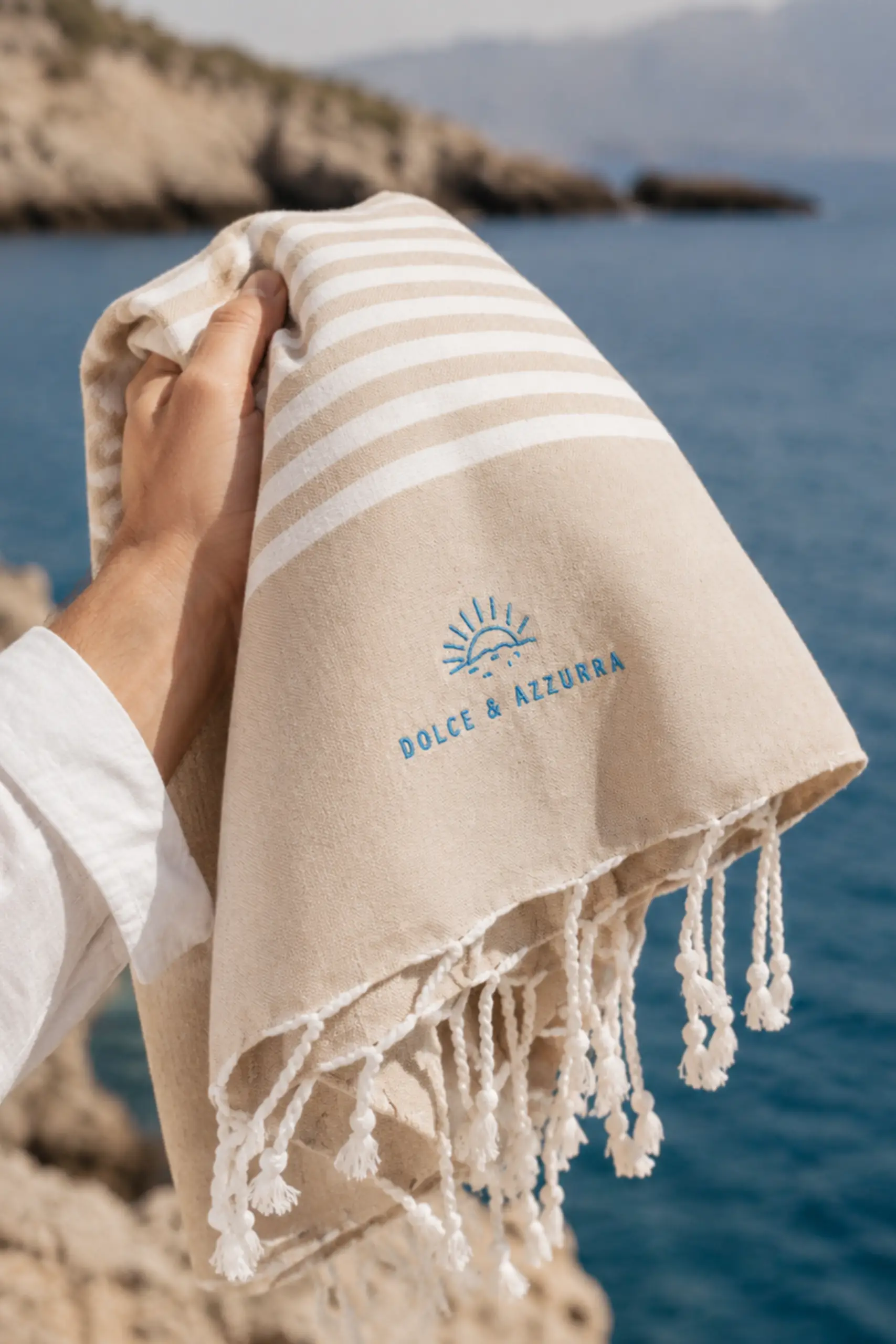 A hand holding a beige beach towel with white stripes, knotted fringing and the embroidered "Dolce & Azzurra" logo, with the sea and rocky coastline in the background.
