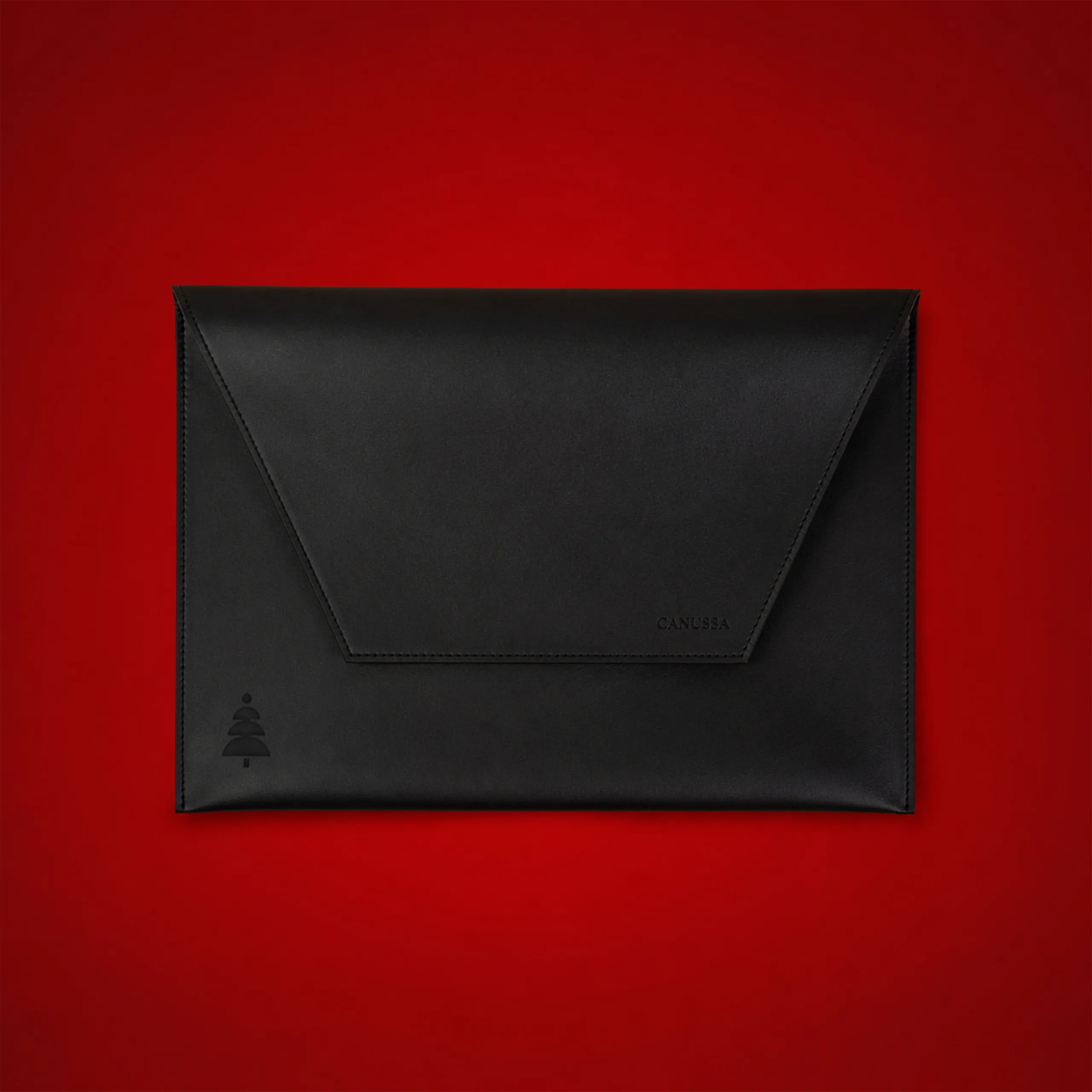 Laptop sleeve made from corn waste in black, with an envelope-style flap and the "Canussa" logo alongside a small embossed Christmas tree, against a red backdrop.