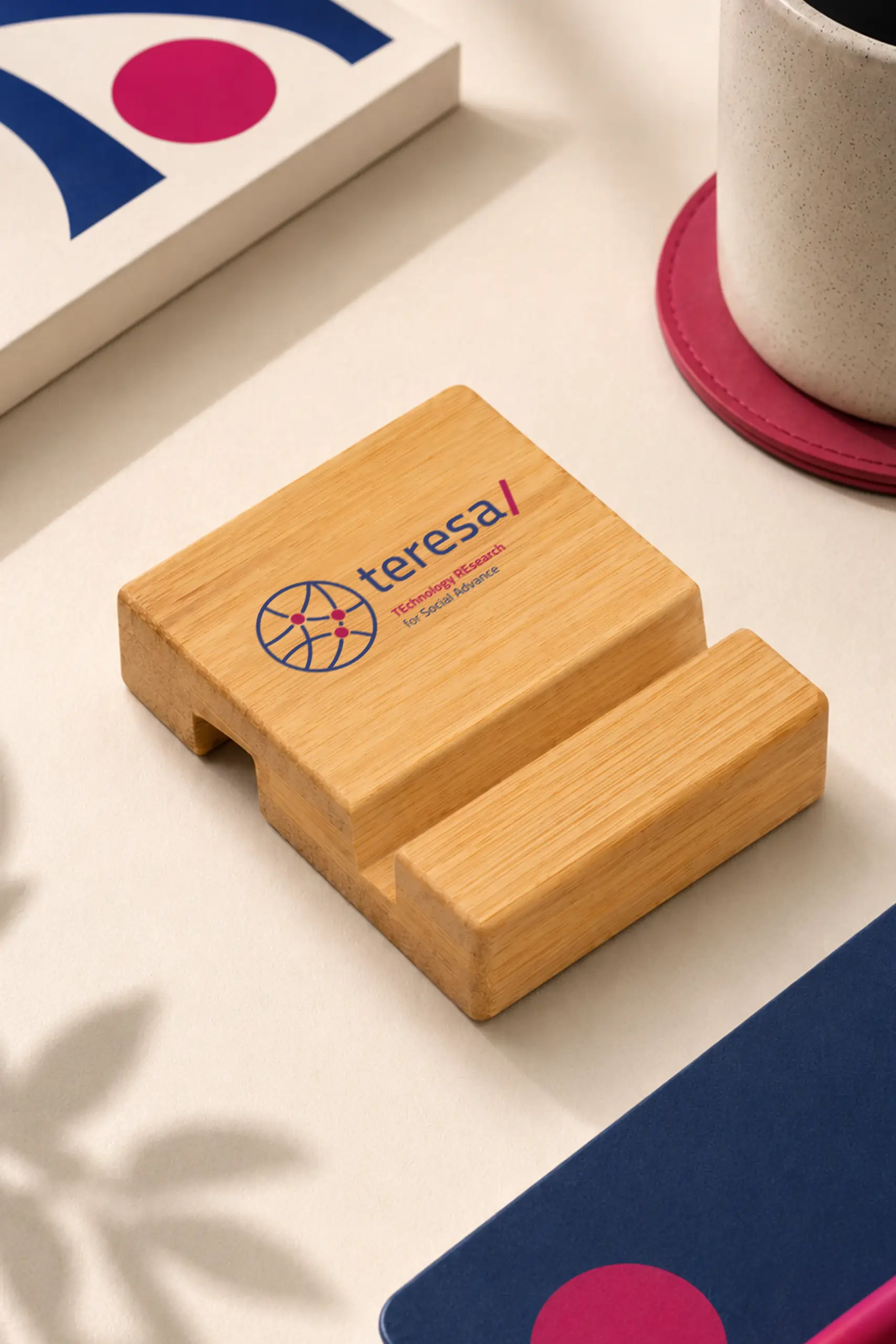Soporte de madera para móvil con el logo "teresa/ TEchnology REsearch for Social Advance" grabado, colocado en un escritorio junto a una taza de cerámica y elementos decorativos en tonos rosa y azul marino.
