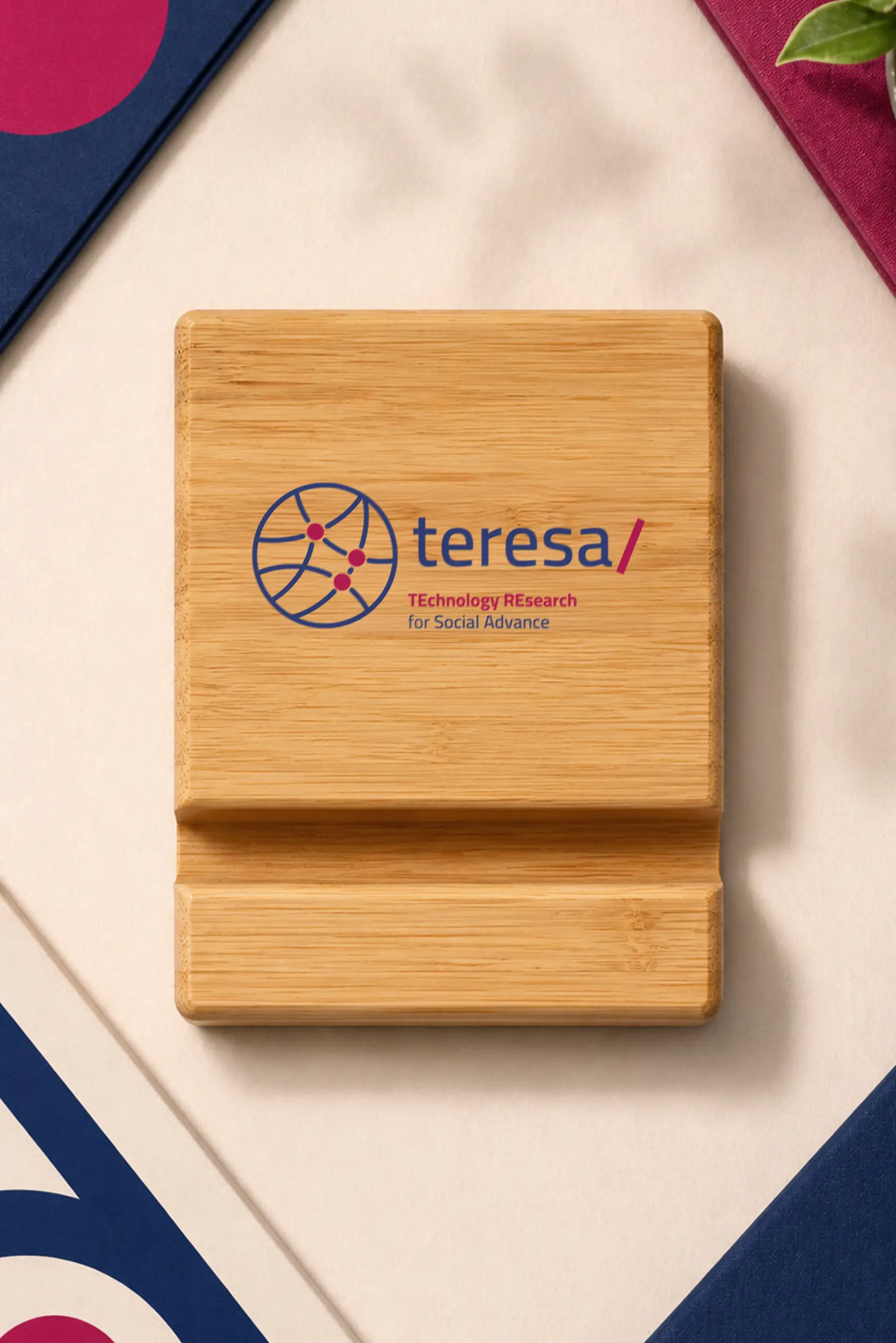 Vista superior de un soporte de madera para móvil con el logo "teresa/ TEchnology REsearch for Social Advance" grabado, sobre una superficie clara con detalles decorativos en rosa y azul.