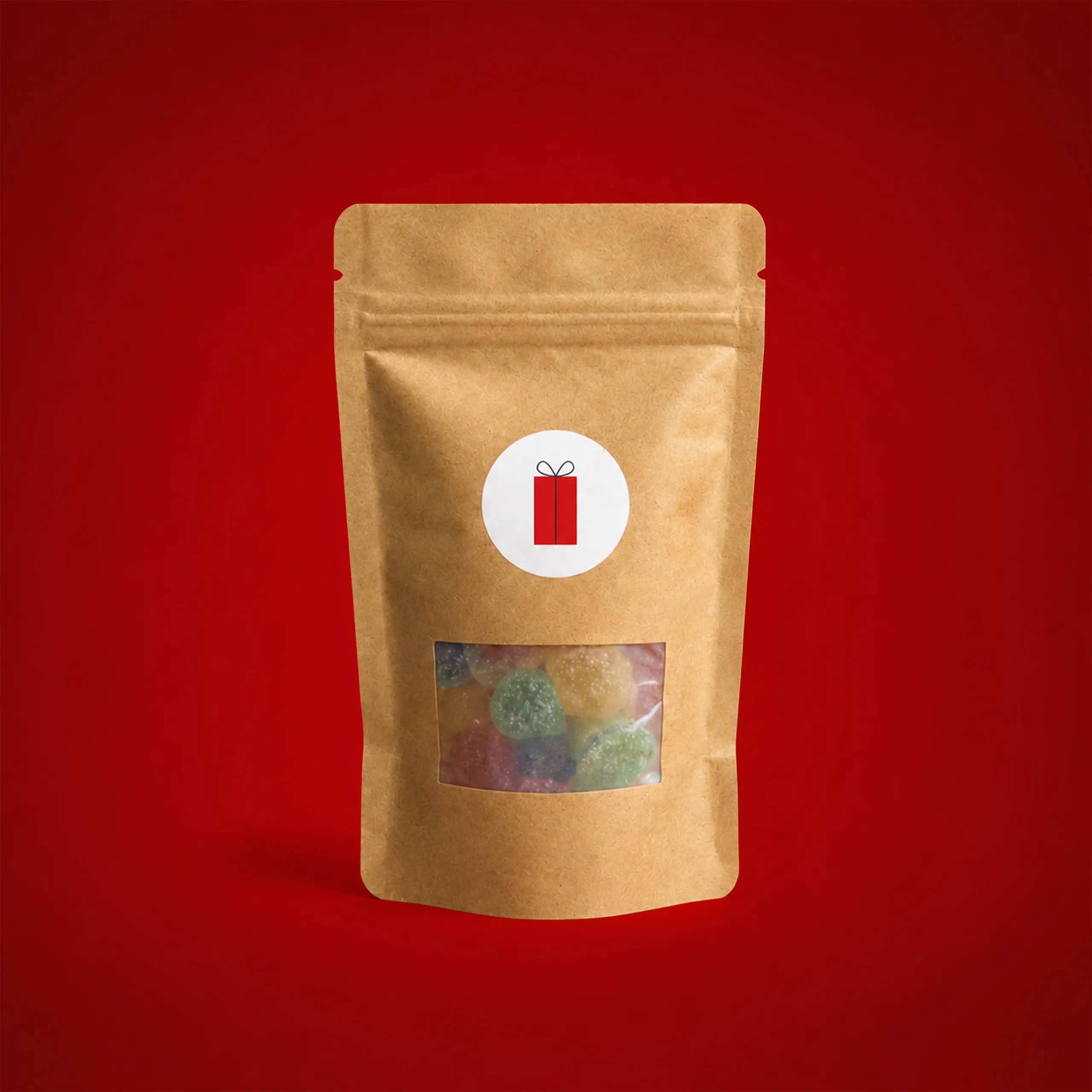 Gominolas personalizadas en bolsa kraft_KRAFTY Bolsa kraft con gominolas personalizada, con ventana troquelada mostrando el contenido y una pegatina circular con la ilustración de un regalo envuelto en rojo, sobre fondo rojo.