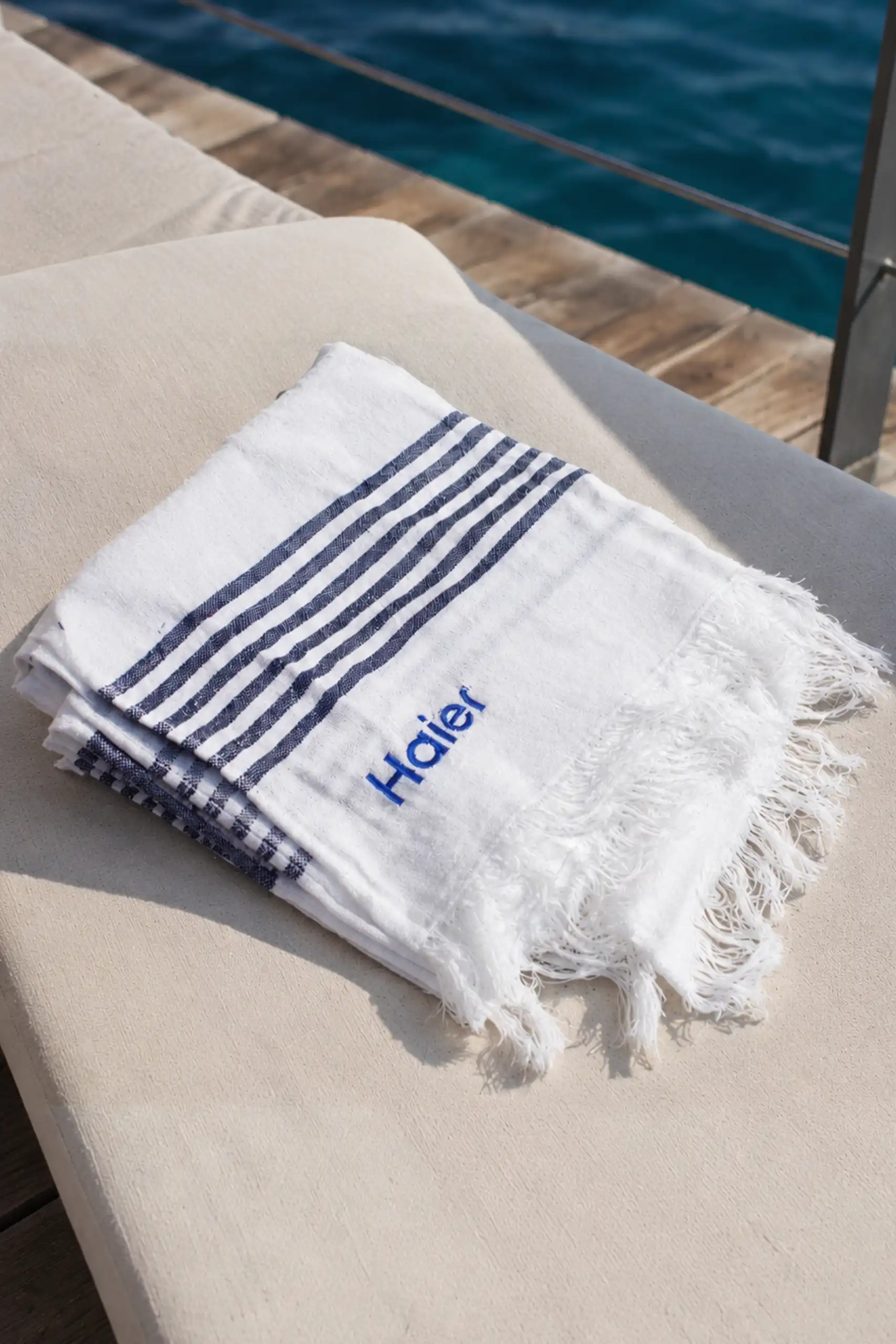 Toalla de playa blanca con rayas azul marino y el logo bordado "Haier", doblada sobre una tumbona junto a una piscina con el mar de fondo.