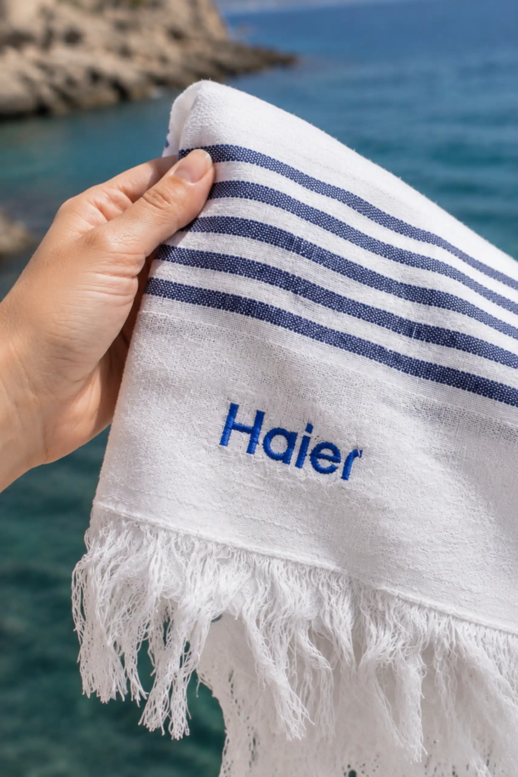 Primer plano de una mano sosteniendo una toalla blanca con rayas azul marino y el logo bordado "Haier", con el mar de fondo.
