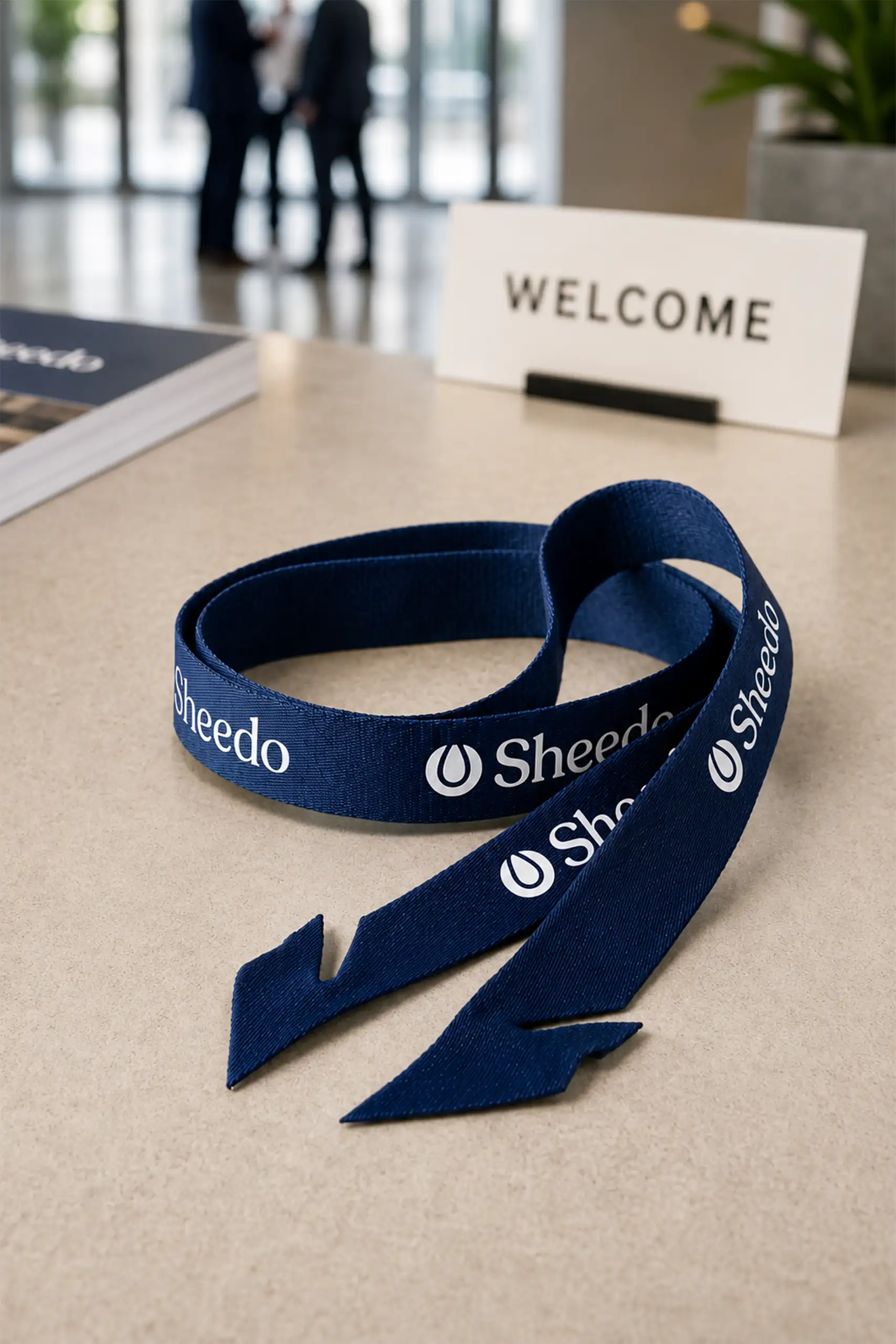 Lanyard rpet sin mosquetón MOSQ Lanyard de rPET con logo Sheedo enrollado sobre un mostrador de recepción, junto a un cartel de bienvenida.