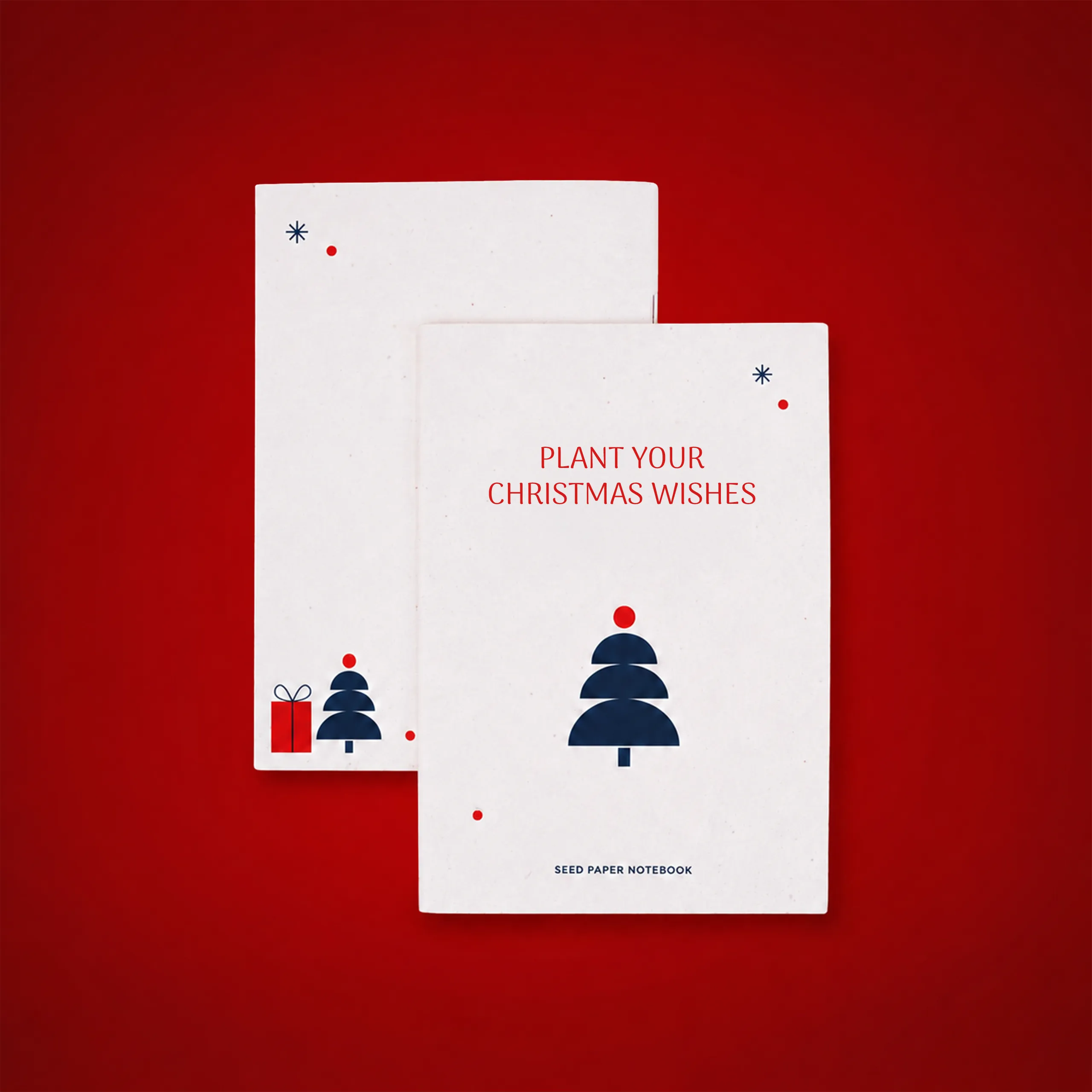 Dos libretas ecológicas con portada de papel plantable, una mostrando el interior en blanco con una ilustración de un regalo y un árbol de Navidad, y la otra con el texto "Plant your Christmas wishes" y "Seed paper notebook" en la portada, sobre fondo rojo.