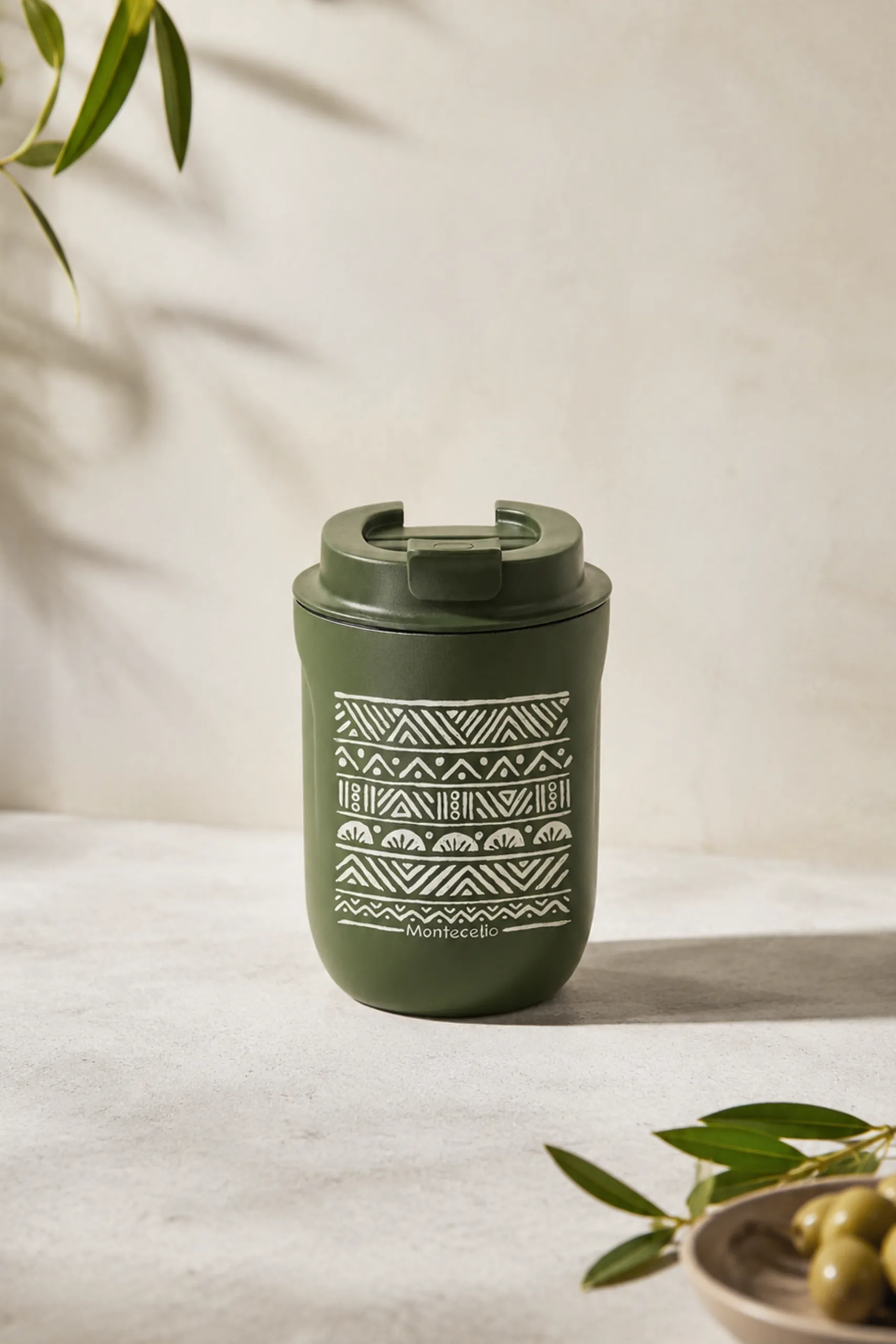 Gobelet thermique réutilisable de couleur vert olive avec un motif géométrique ethnique blanc et le texte « Montecelio », posé sur une surface claire aux côtés de branches d'olivier.