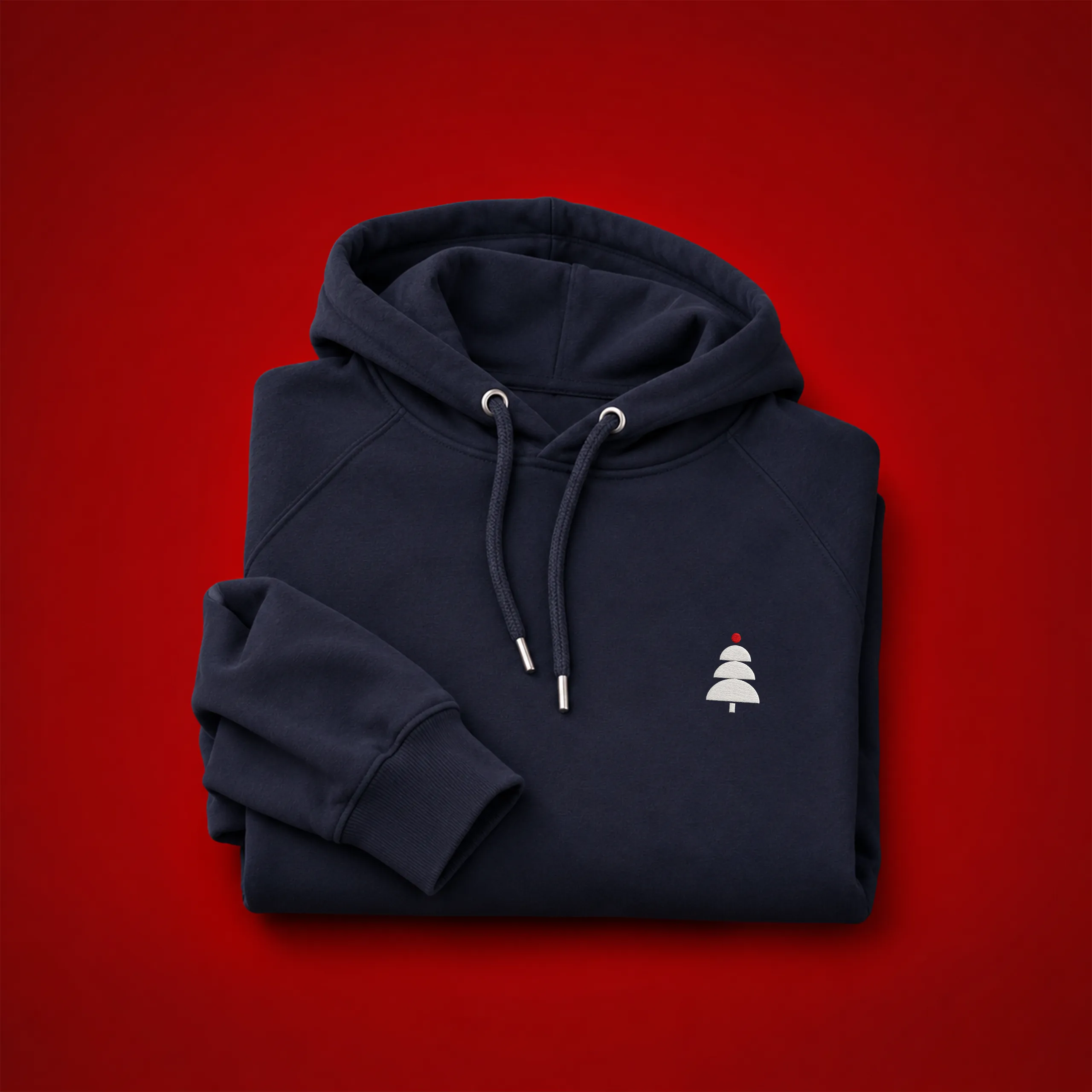 Sudadera personalizada con capucha_HUVA Sudadera con capucha personalizada en azul marino, doblada y mostrando un pequeño árbol de Navidad bordado en blanco con una bola roja, sobre fondo rojo.