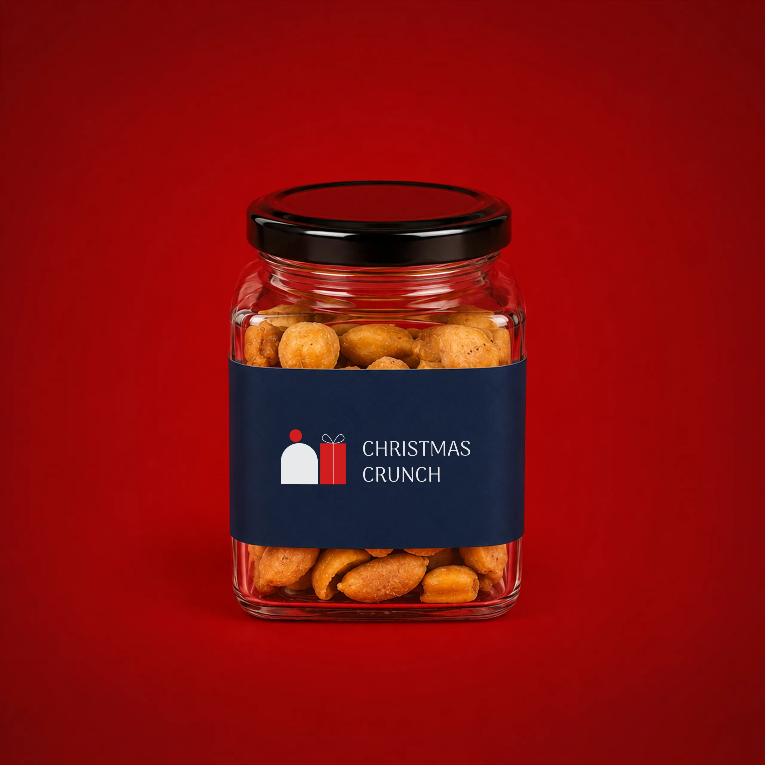 Tarro de cristal con mix de frutos secos_NUTSY Tarro de cristal con mix de frutos secos, con tapa negra y etiqueta azul marino mostrando el texto "Christmas Crunch" junto a una ilustración de una figura y un regalo, sobre fondo rojo.