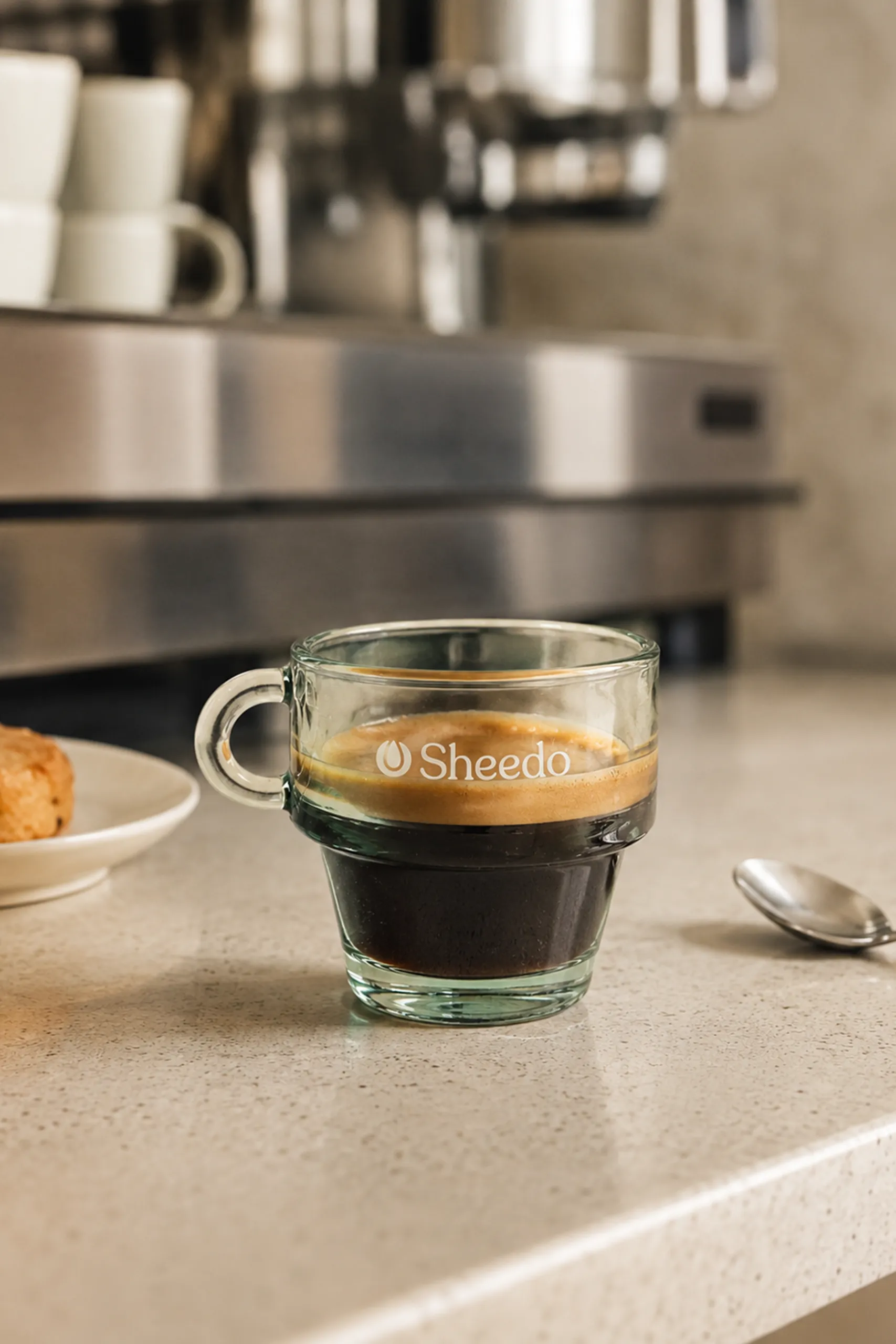 Recycled glass cup with Sheedo logo filled with espresso coffee on a worktop, next to a teaspoon and a plate of pastries.