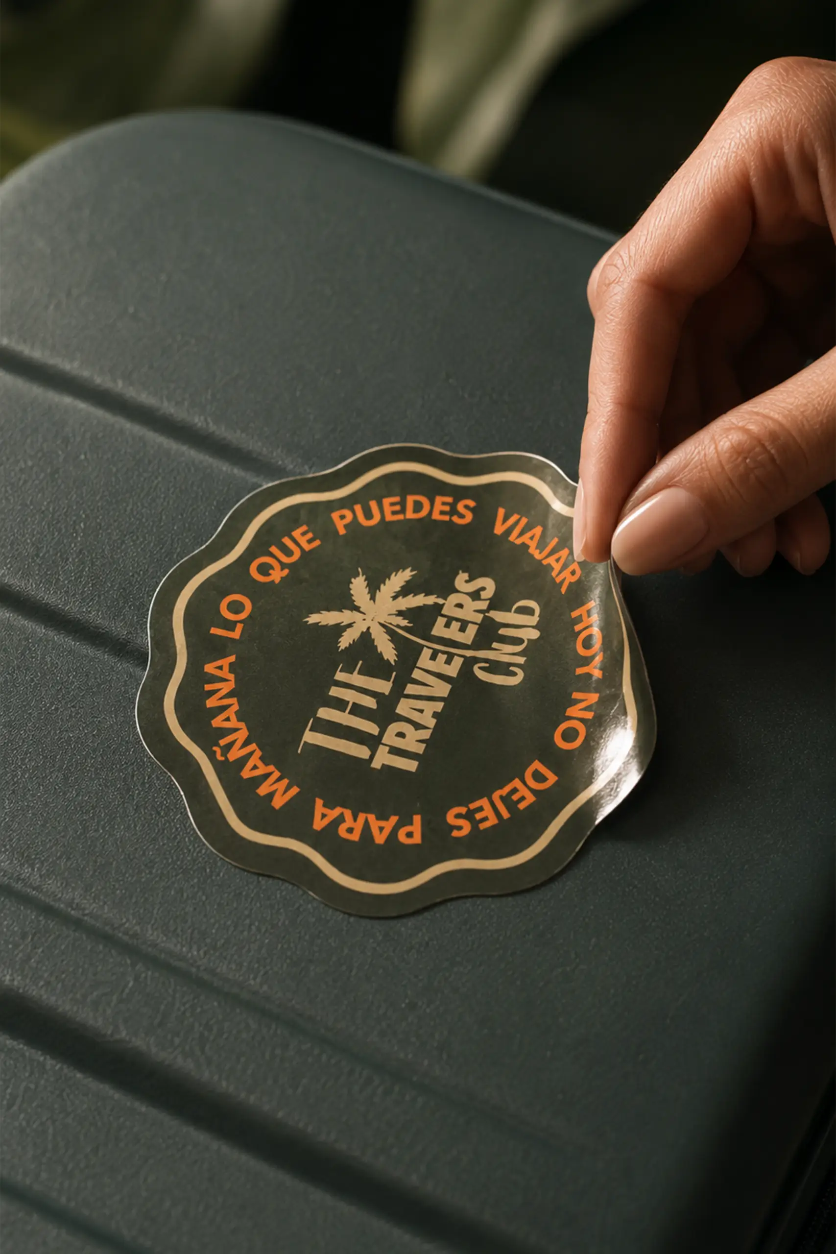 Mano colocando una pegatina troquelada de color verde personalizada con la imagen de la marca "The Trevelers Club" sobre una maleta de viaje verde