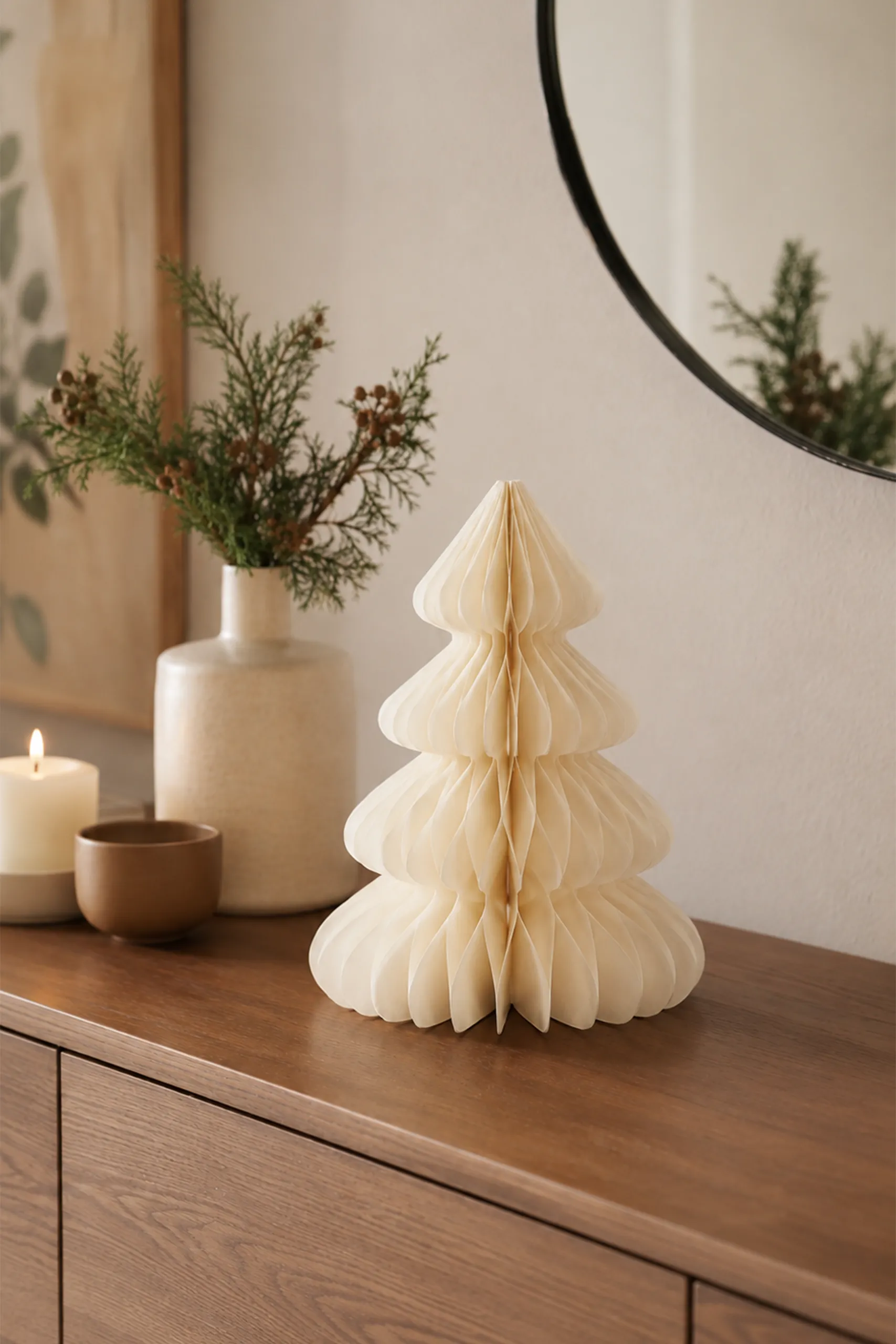 Árbol de Navidad de papel plegable en color crudo colocado sobre un mueble de madera, junto a un jarrón con ramas de eucalipto, una vela encendida y un cuenco, con un espejo redondo reflejando la decoración.