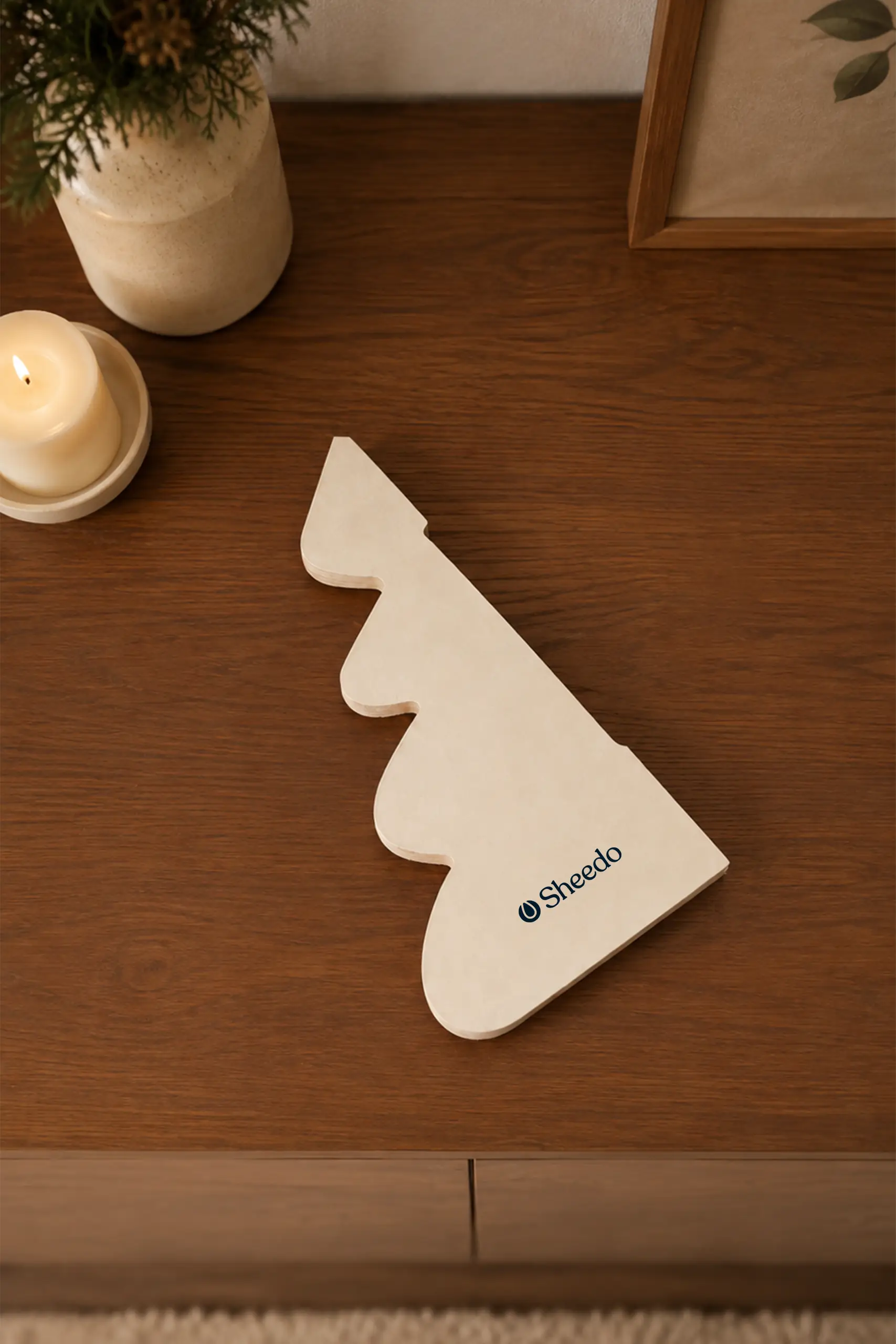 Pieza plana en forma de árbol de Navidad en color crudo con el logo "Sheedo" impreso, colocada sobre un mueble de madera junto a un jarrón y una vela encendida, antes de su montaje.