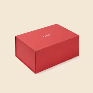 Caja de regalo roja con cierre magnético, vista en ángulo sobre fondo beige, con el logotipo "L*GO" grabado en la tapa.