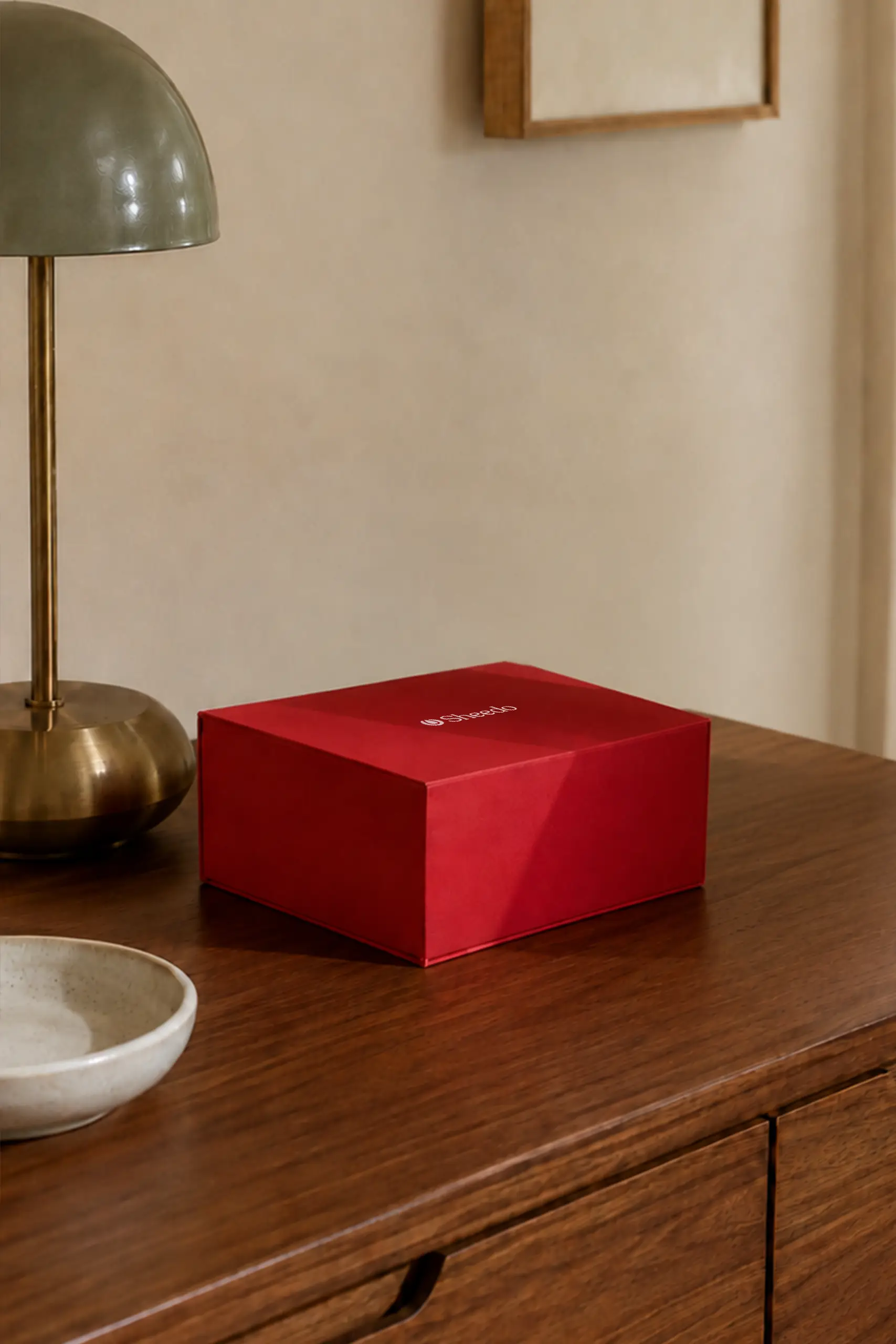Caja de regalo roja con el logotipo "Sheedo" grabado, vista desde arriba sobre un mueble de madera oscura junto a una lámpara dorada, un cuenco de cerámica y un cuadro en la pared.