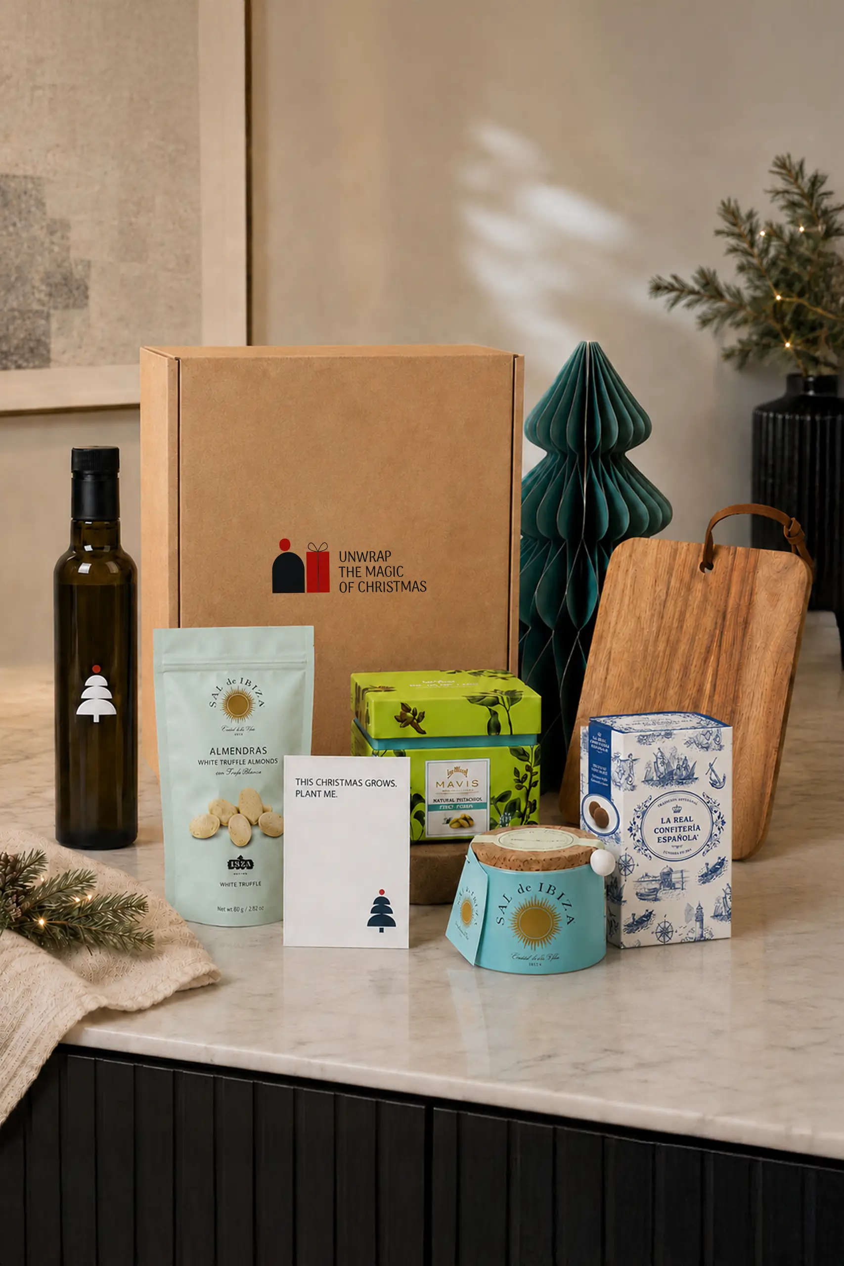 "Premium" Christmas hamper featuring a kraft box printed with the "Unwrap the magic of Christmas" logo, a bottle of olive oil, a pack of Sal de Ibiza white truffle almonds, a decorative tin of Mavis natural pistachios, a wooden board with a leather handle, a box of La Real Confitería Española nougat, a Sal de Ibiza salt container, a plantable card reading "This Christmas grows. Plant me.", and a decorative teal paper Christmas tree, arranged on a kitchen counter with festive decorations in the background.