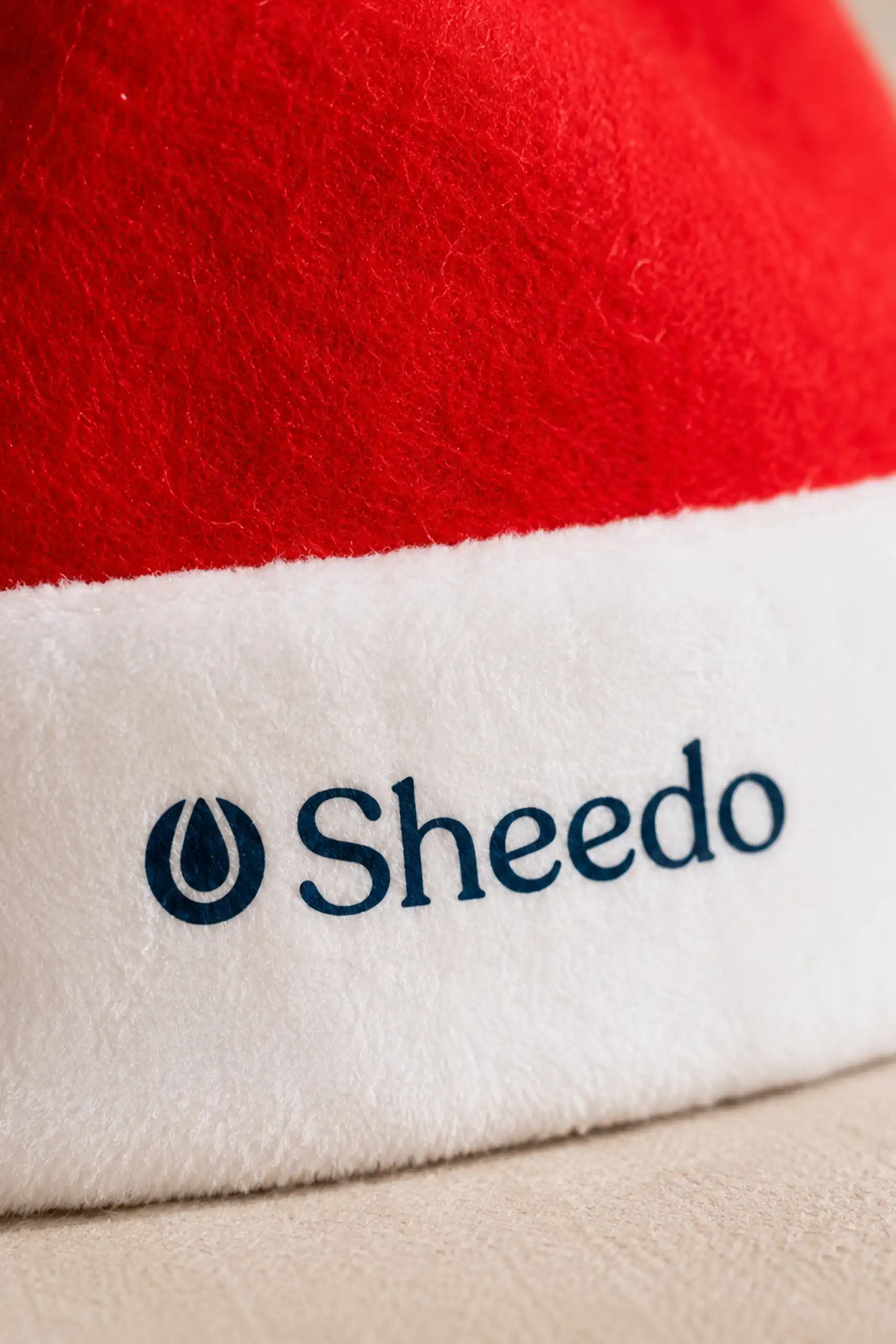 Primer plano del logotipo "Sheedo" bordado en el borde blanco del gorro de Papá Noel, mostrando el detalle del bordado y la textura de la tela.