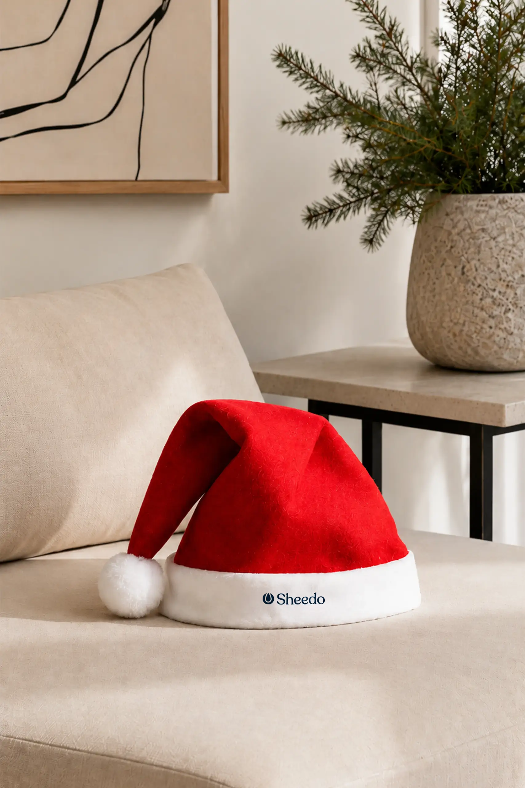 Gorro de Papá Noel personalizado con logotipo "Sheedo" bordado en el borde blanco, colocado sobre un sofá beige en un ambiente navideño con un jarrón y ramas de pino.