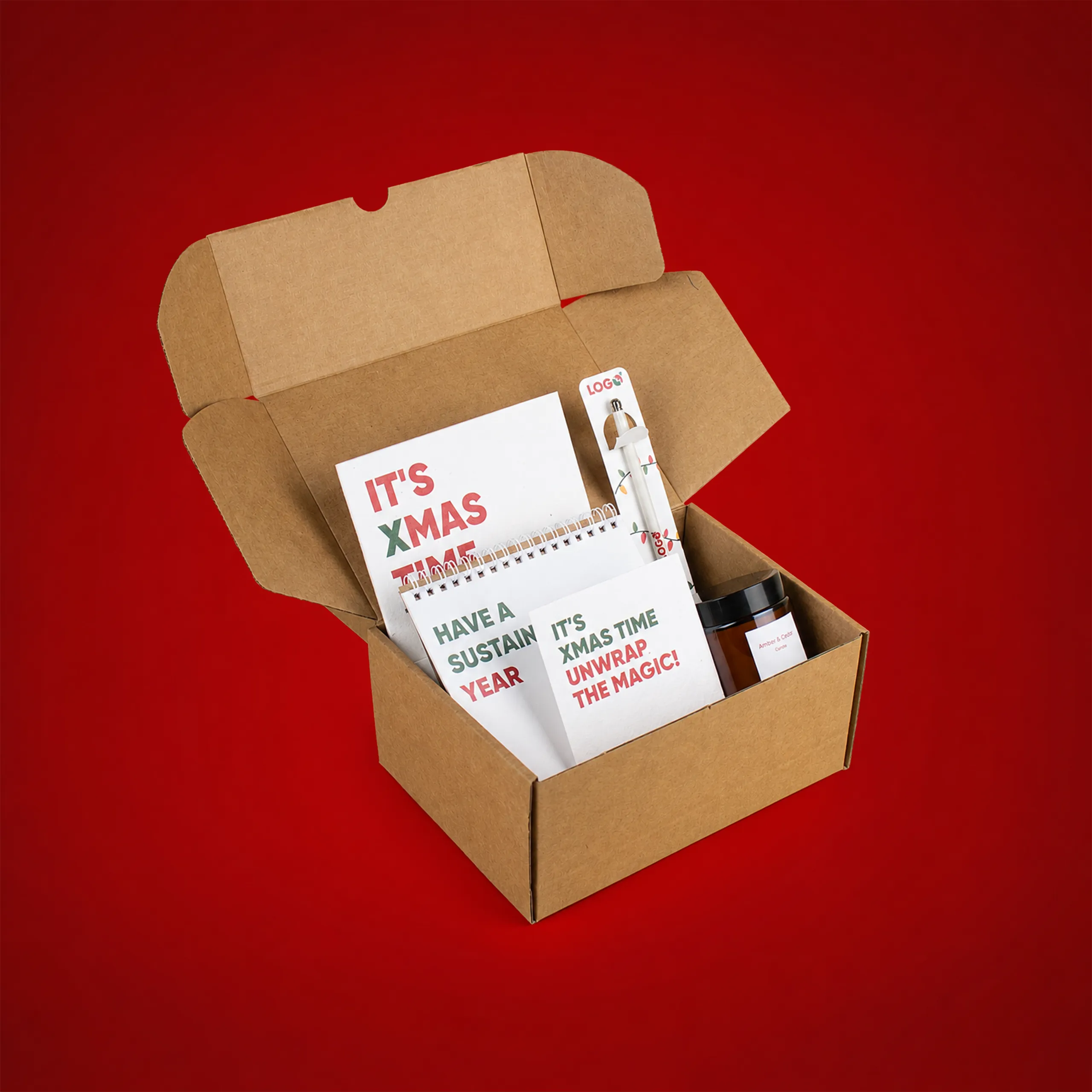 Open kraft cardboard box containing a Christmas gift pack, including a spiral-bound notebook reading "Have a sustainable year", cards printed with the messages "It's Xmas Time" and "Unwrap the magic!", a pen decorated with festive fairy lights, and an amber glass jar candle scented "Amber & Cedar", set against a red background.