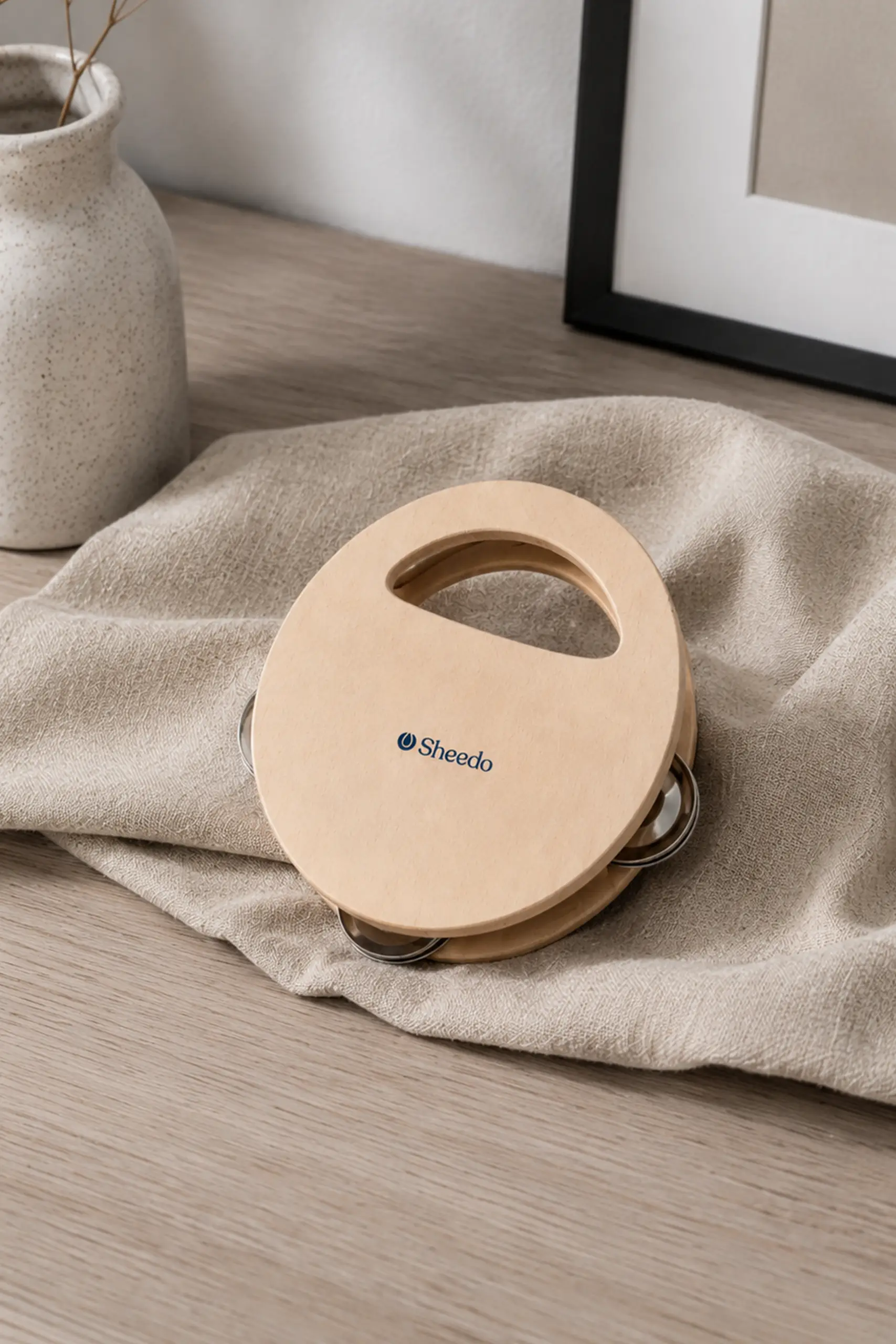 Pandereta de madera personalizada con el logotipo "Sheedo", colocada sobre un paño de lino beige en una composición de estilo de vida con jarrón de cerámica y marco de fotos al fondo.