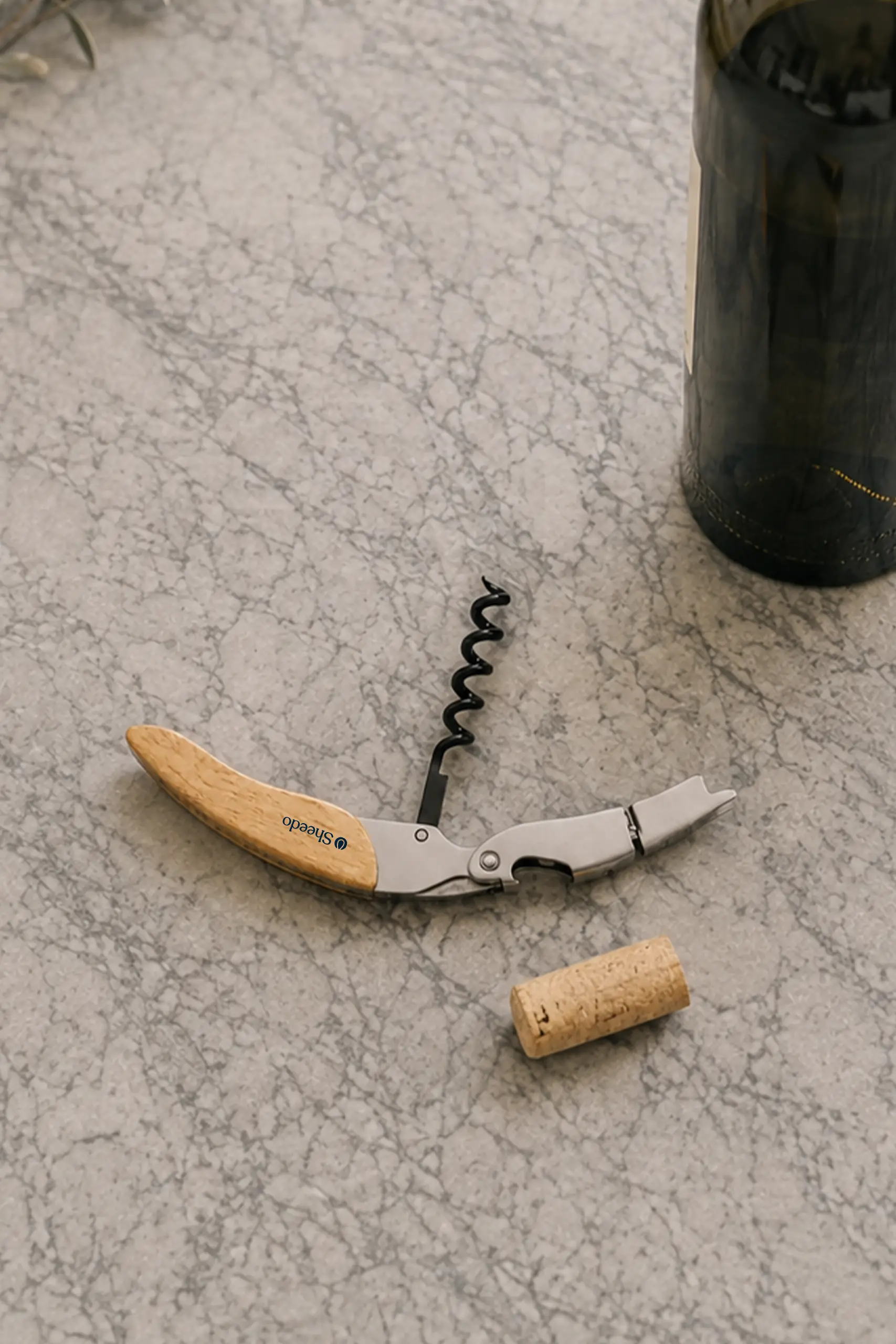 Sacacorchos de madera con el logotipo "Sheedo" grabado, apoyado sobre una encimera de mármol junto a un corcho extraído y una botella de vino.