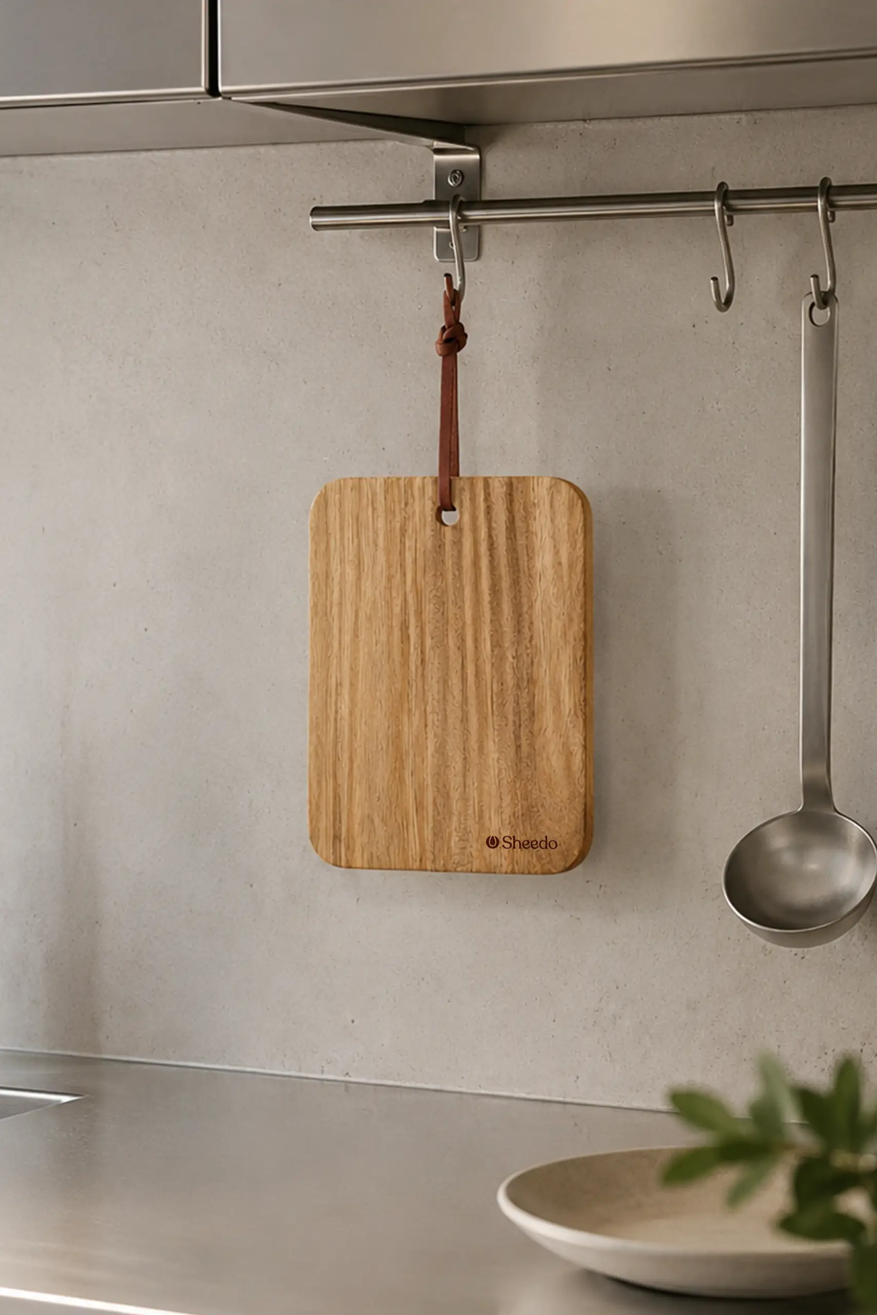Tabla de cortar de madera_PARMA Tabla de cortar de madera con cordón de cuero sobre una encimera de acero inoxidable, con unas manos cortando una aceituna con un cuchillo, en una cocina con un jarrón de cerámica al fondo.