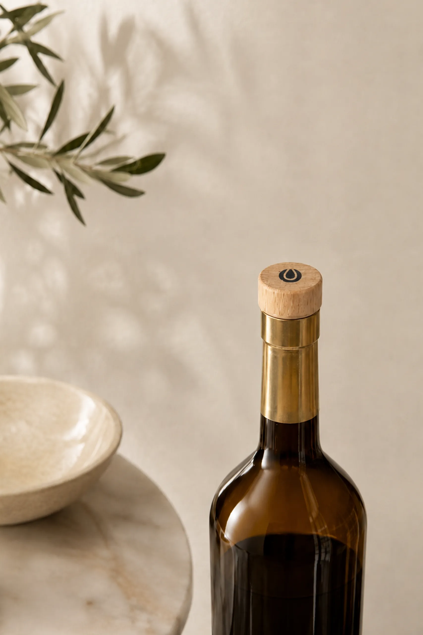 Reusable wooden bottle stopper with an engraved finish, fitted on the neck of an amber glass bottle, in a decorative setting with an olive branch and a ceramic bowl.