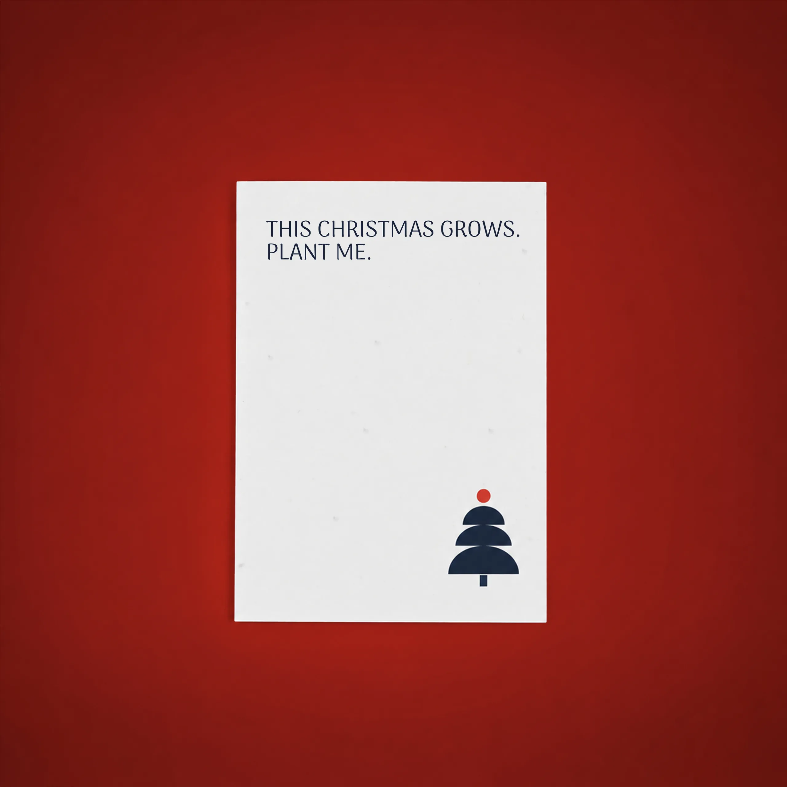 Carte de Noël plantable de couleur blanche portant le texte « This Christmas grows. Plant me. », ornée d'un petit sapin de Noël bleu marine surmonté d'une boule rouge dans le coin inférieur droit, sur fond rouge.