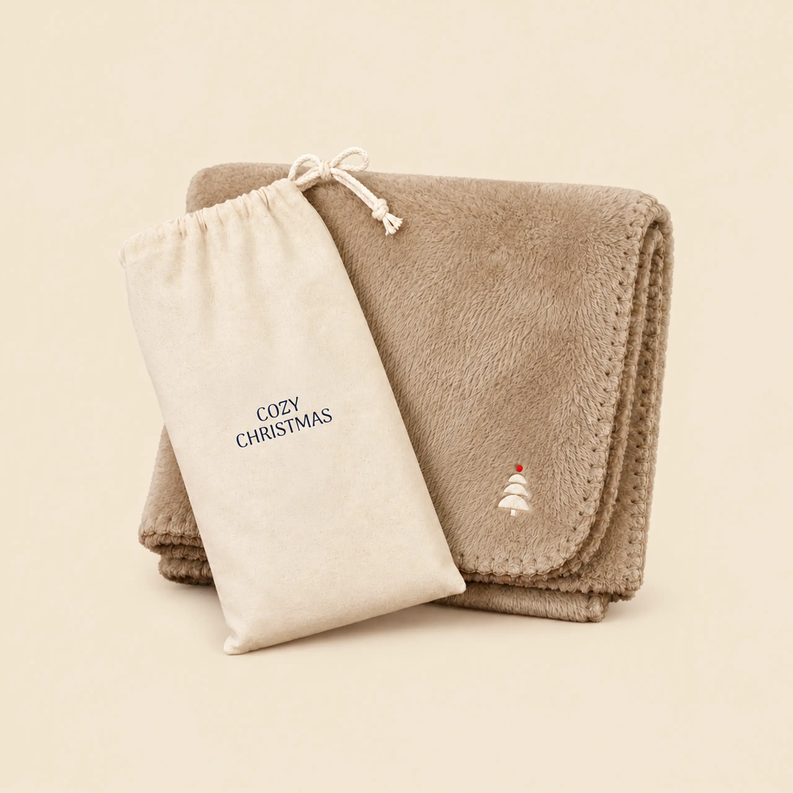 Manta polar beige doblada con árbol bordado y bolsa de algodón con cordón y el texto «Cozy Christmas».