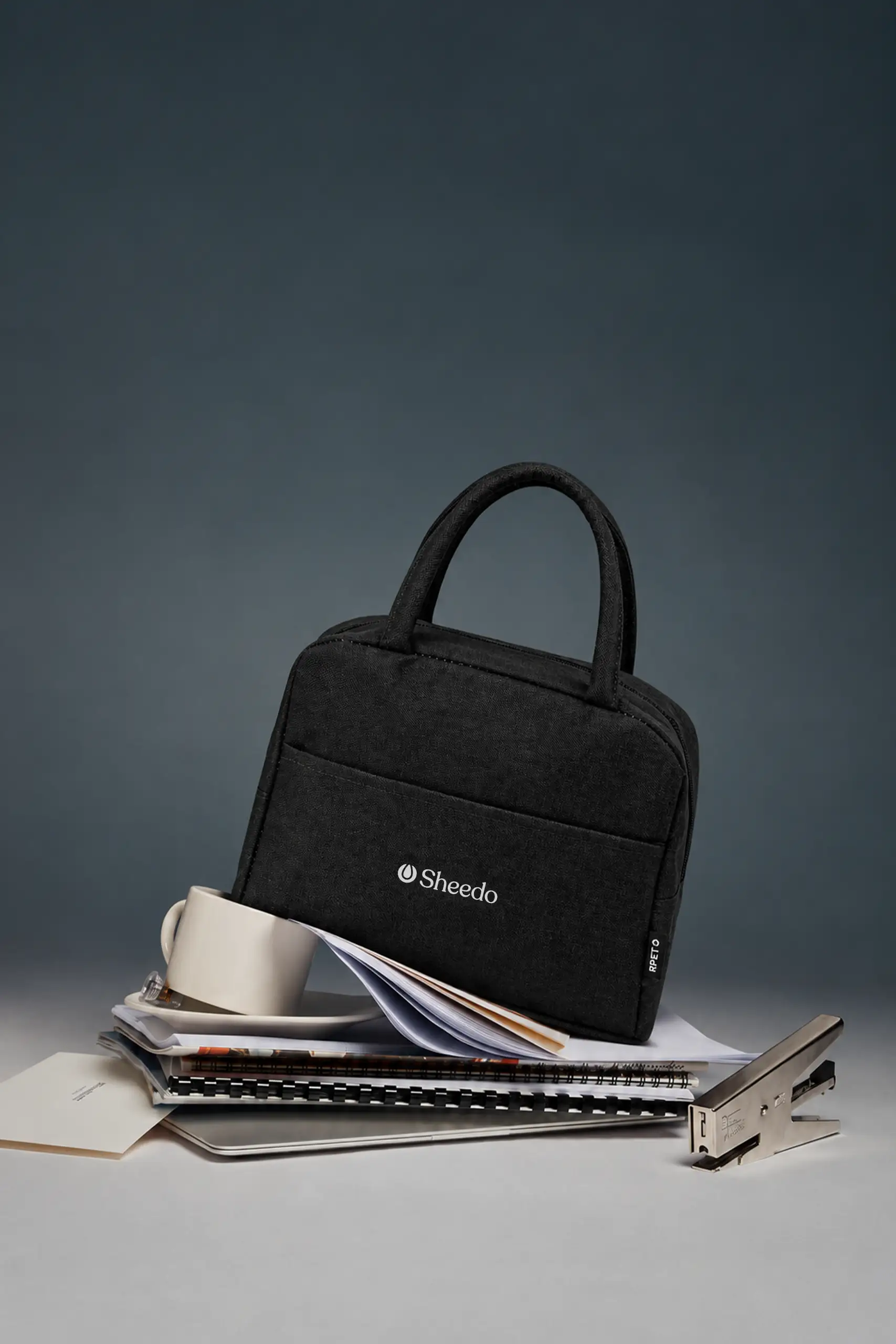 Bolsa nevera rPET negra para llevar la comida al trabajo, con logo de Sheedo, junto a documentos, taza y grapadora