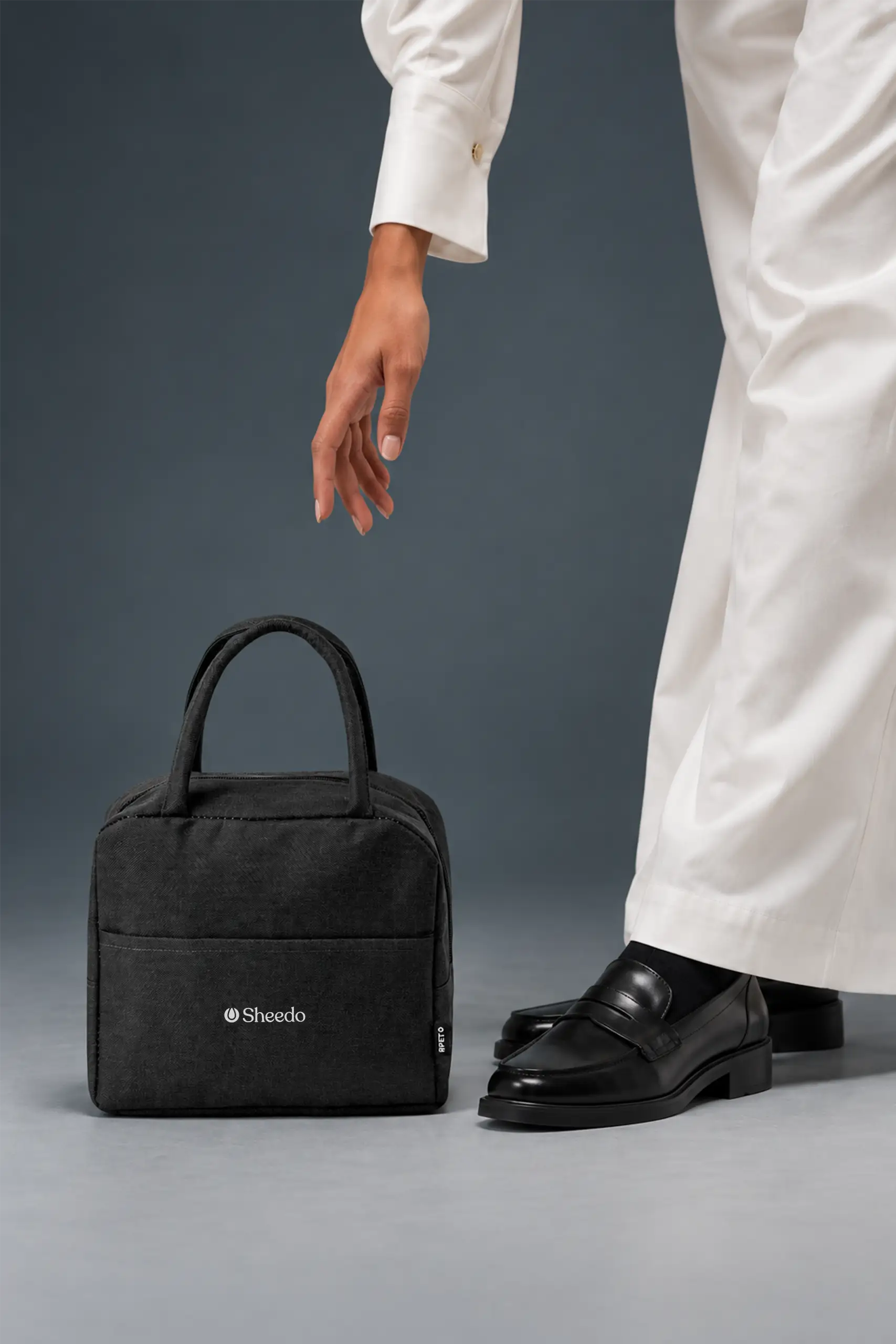 Persona con pantalón blanco y mocasines alargando la mano hacia una bolsa nevera rPET negra con logo de Sheedo