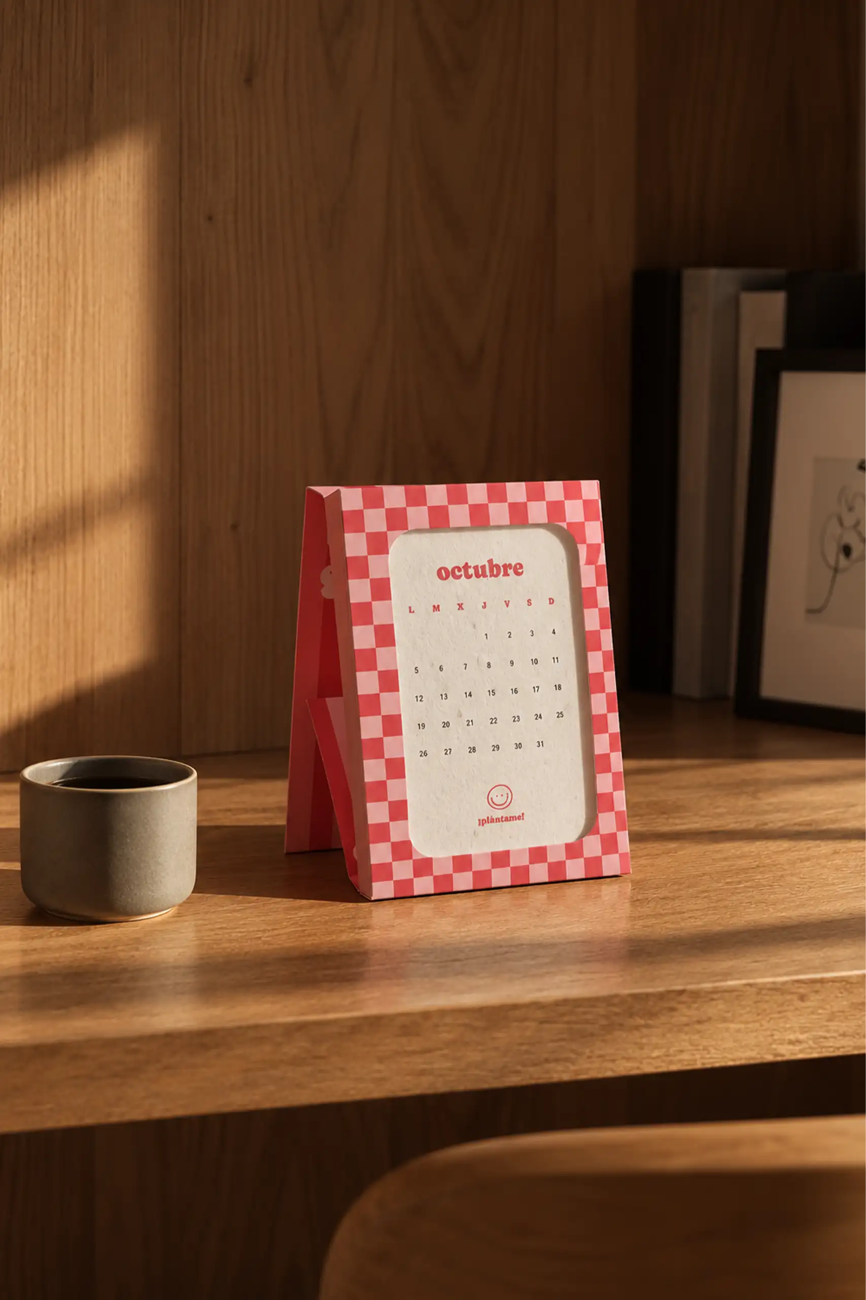 Calendario plantable personalizado para Nanuc.fun montado como caballete de sobremesa, con marco de cuadros rojos y rosas y la hoja de octubre en papel de semillas con el mensaje 