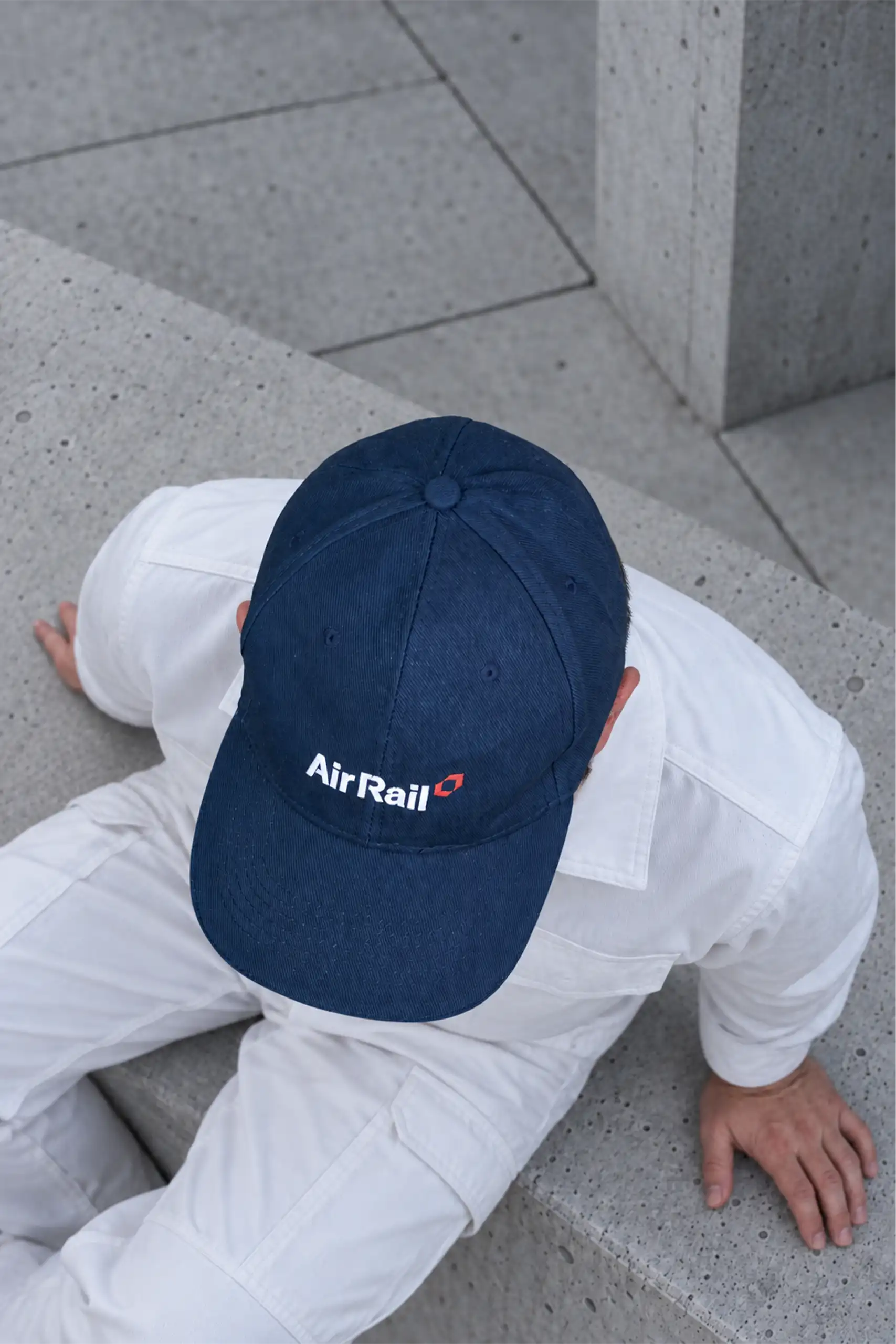 Top view of a person dressed in white sitting on concrete, wearing the navy 100% cotton cap customised for Air Rail with its logo embroidered in white and red.