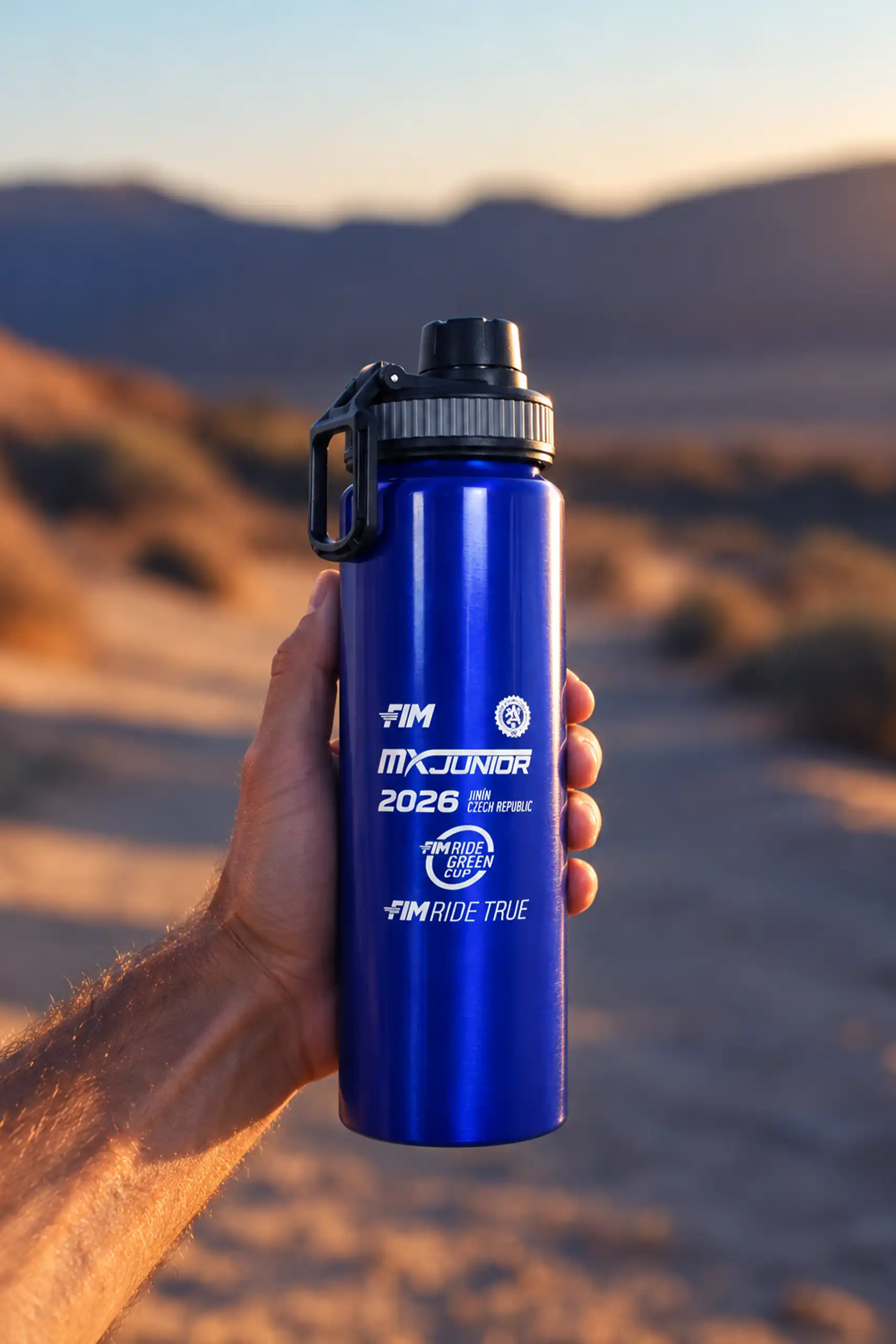 Hand holding the custom FIM sports bottle in blue, printed in white with the MX Junior 2026, FIM Ride Green Cup and FIM Ride True logos, against a desert landscape at sunset.