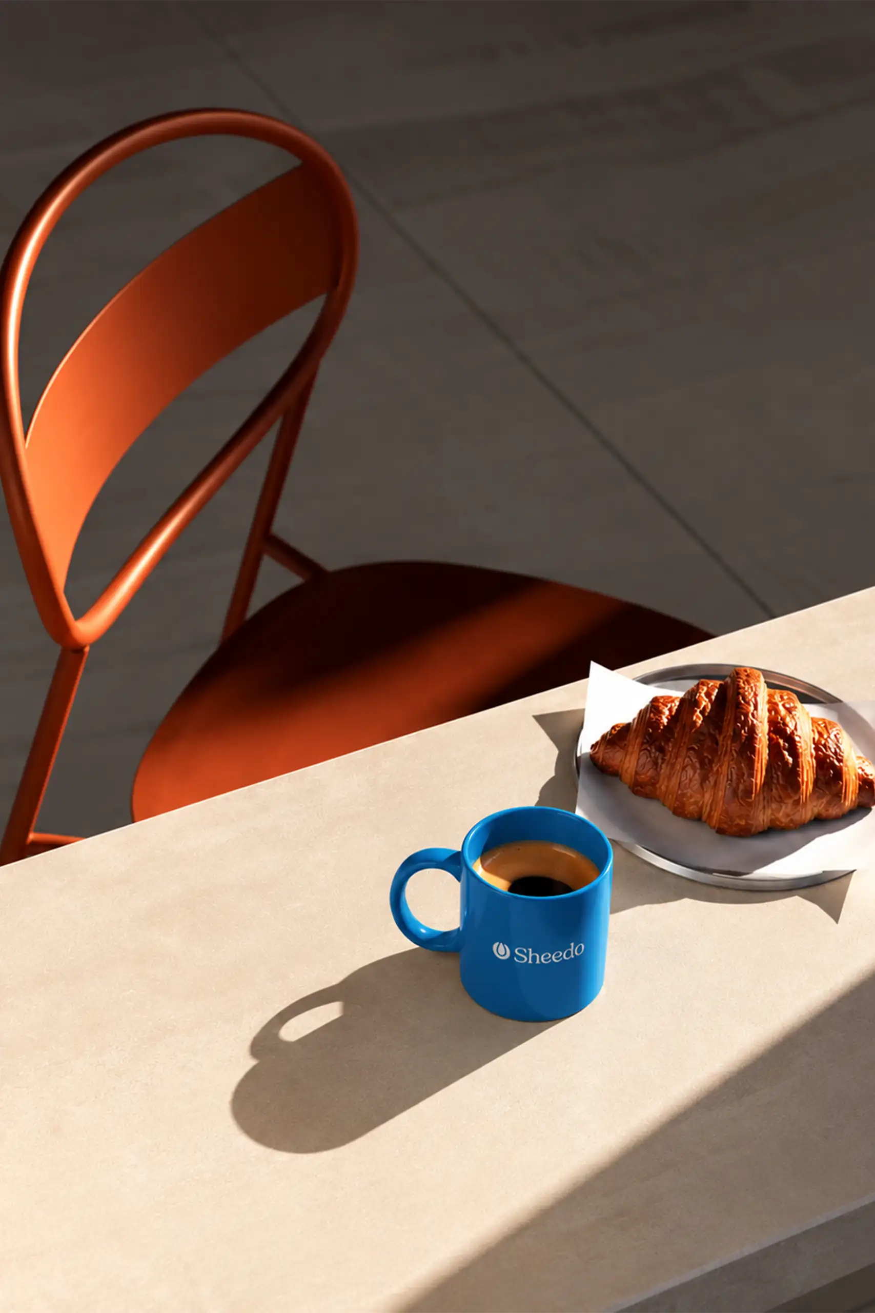 Taza de cerámica azul personalizada con logo Sheedo y café junto a un croissant en una mesa soleada con silla naranja