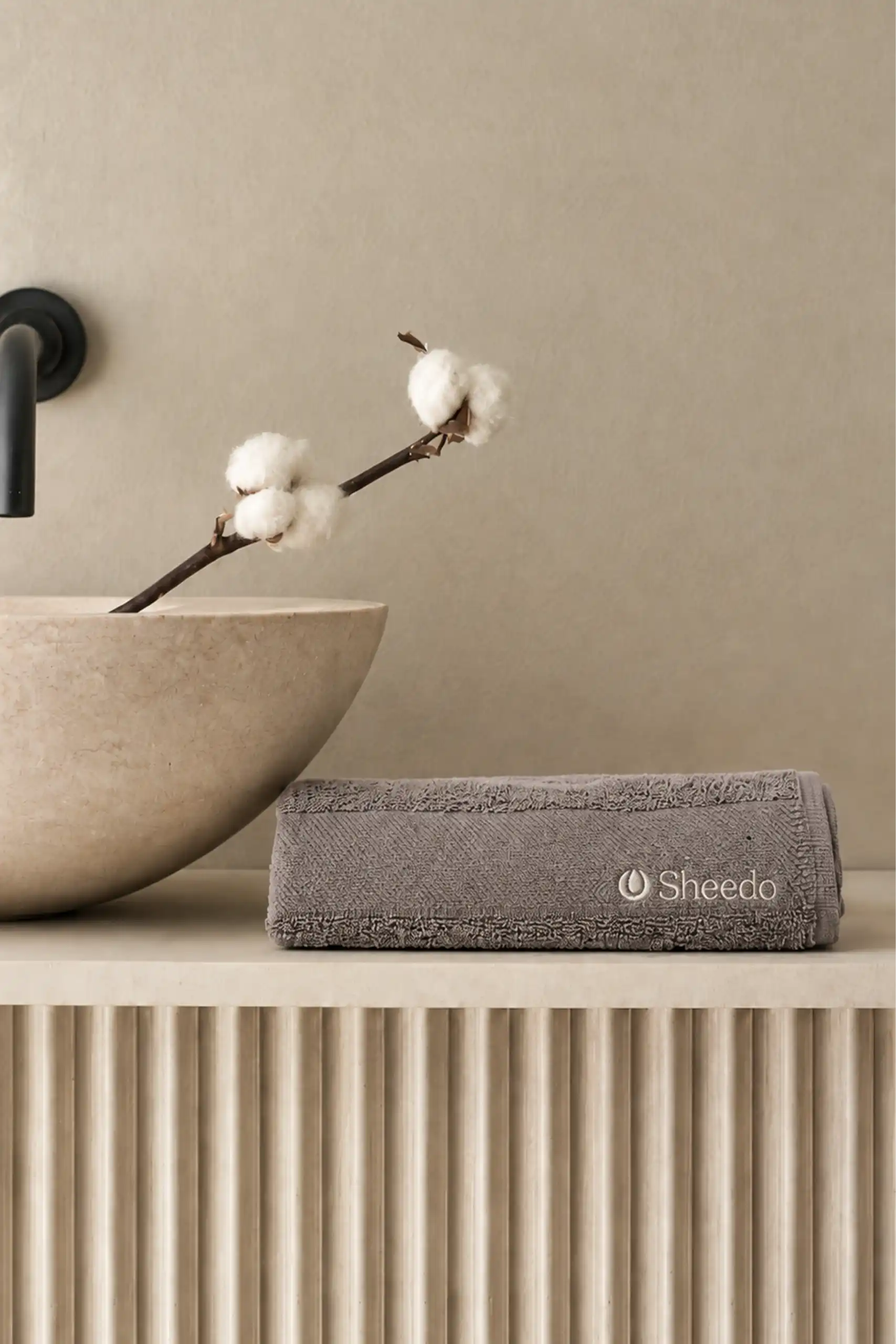 Toalla de manos personalizada CLARA Toalla de manos personalizada gris doblada con logotipo Sheedo bordado, en un baño de tonos beige con lavabo de piedra y flores de algodón